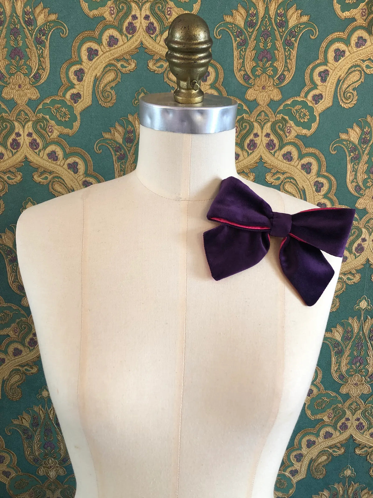 Broochella handmade Small Velvet Bow Brooch in deep purple velvet with contrasting fuchsia lining, styled on the shoulder of a cream mannequin against ornate green and gold wallpaper.