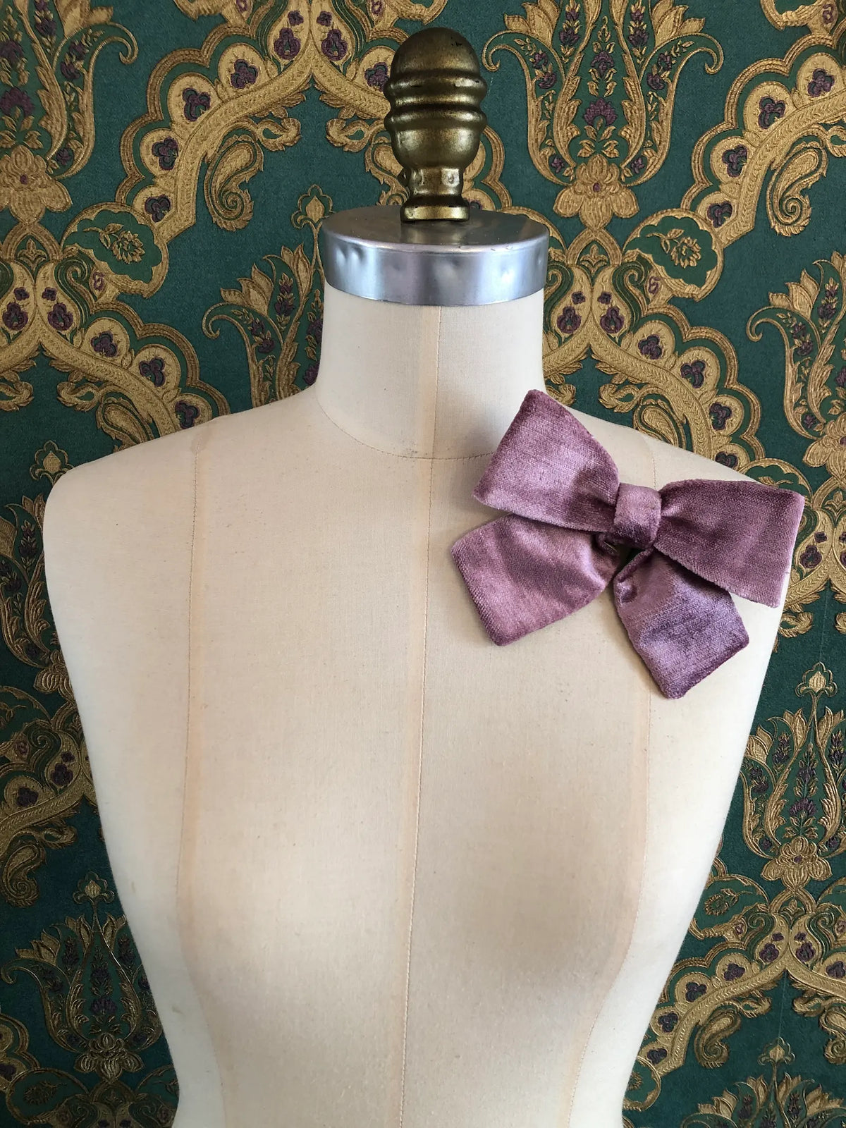 Broochella handmade Small Velvet Bow Brooch in soft violet velvet with contrasting green lining, styled on the shoulder of a cream mannequin against ornate green and gold wallpaper.