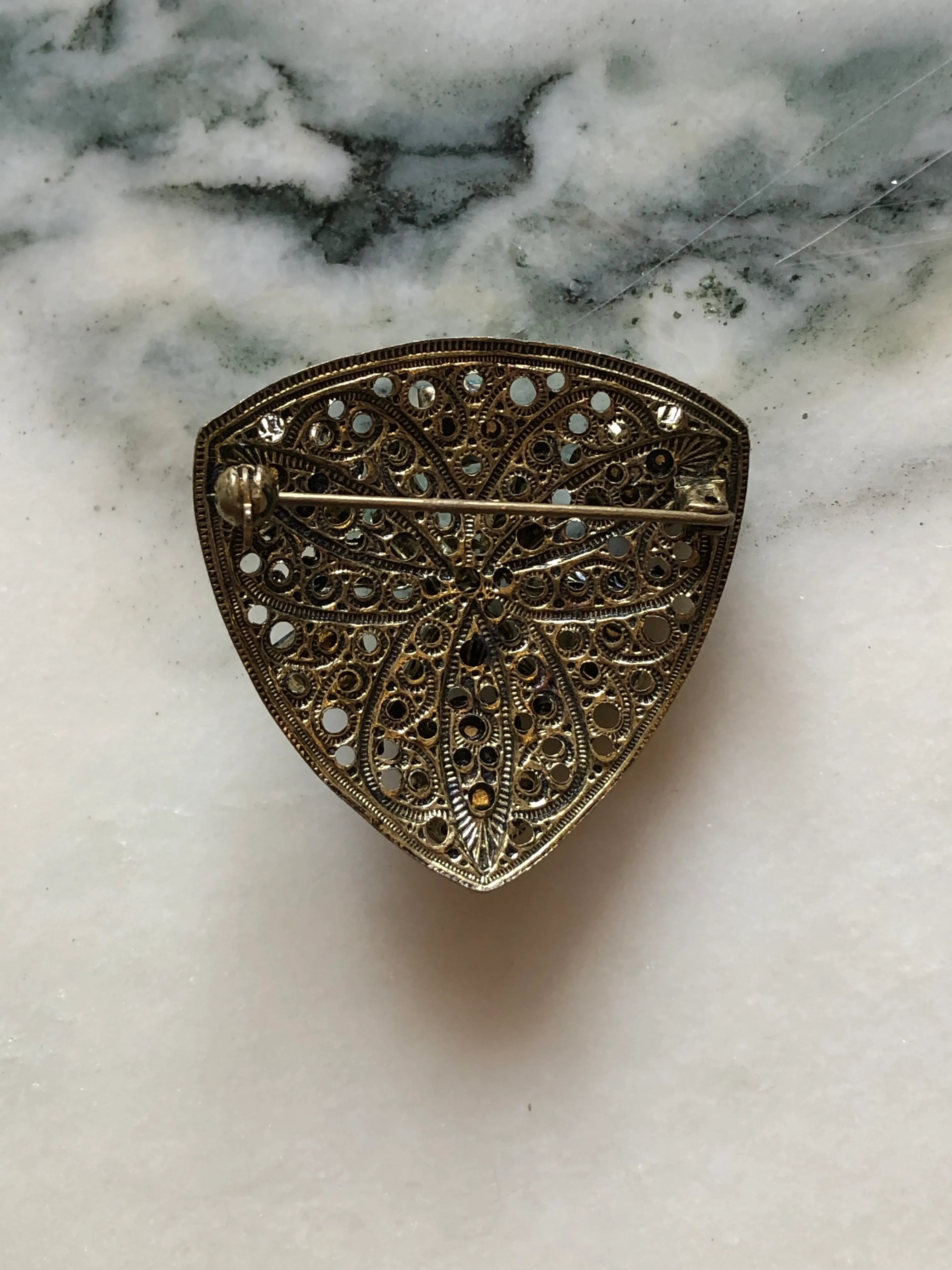 The back view of Broochella Smaraldo vintage Czech shield brooch with emerald-green crystals and ornate antiqued silver-tone filigree. the background is white marble