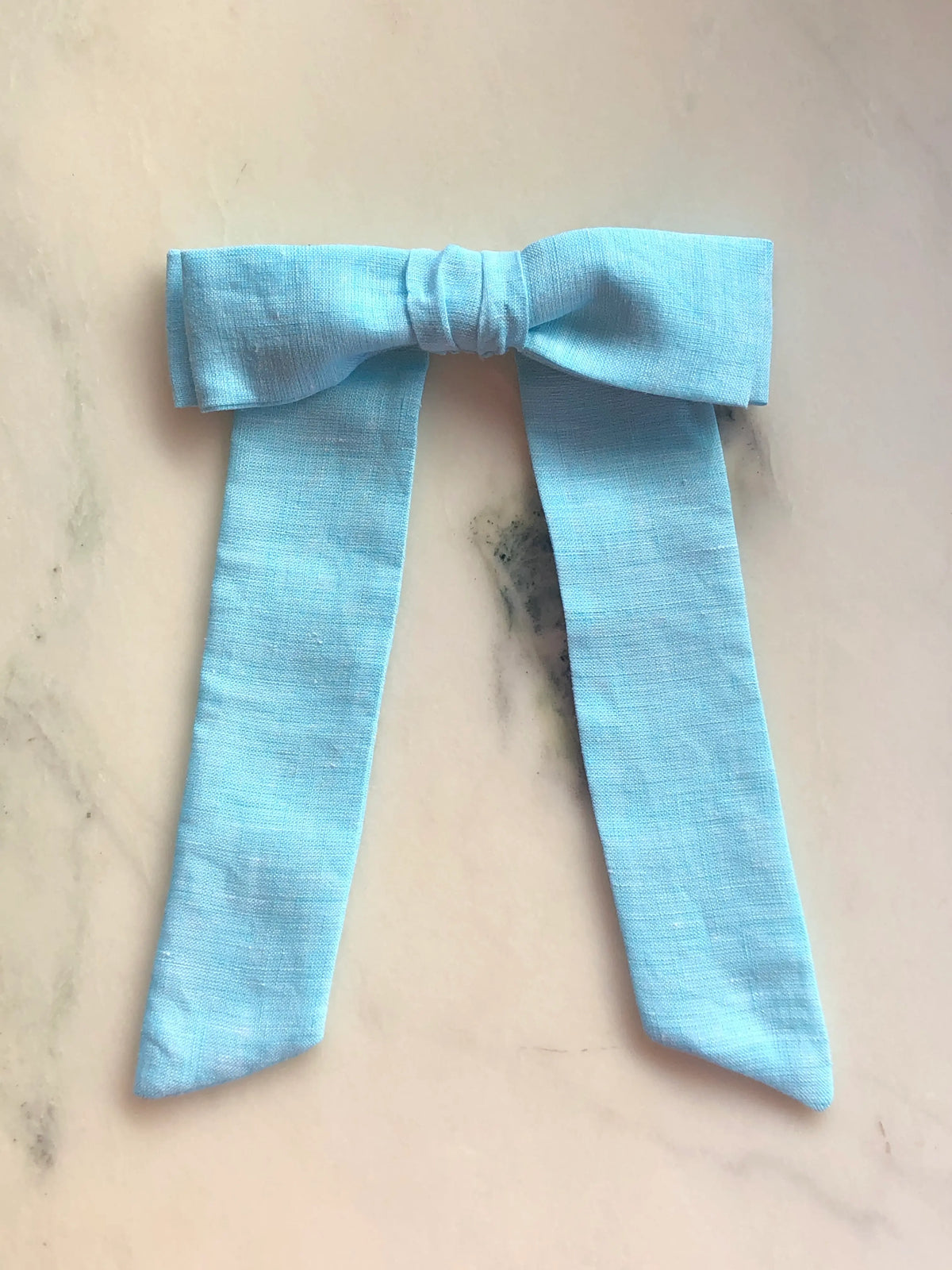Broochella Sofia blue linen bow brooch, a handmade designer statement accessory with an oversized bow and elegant long ribbon tails, photographed on white marble.