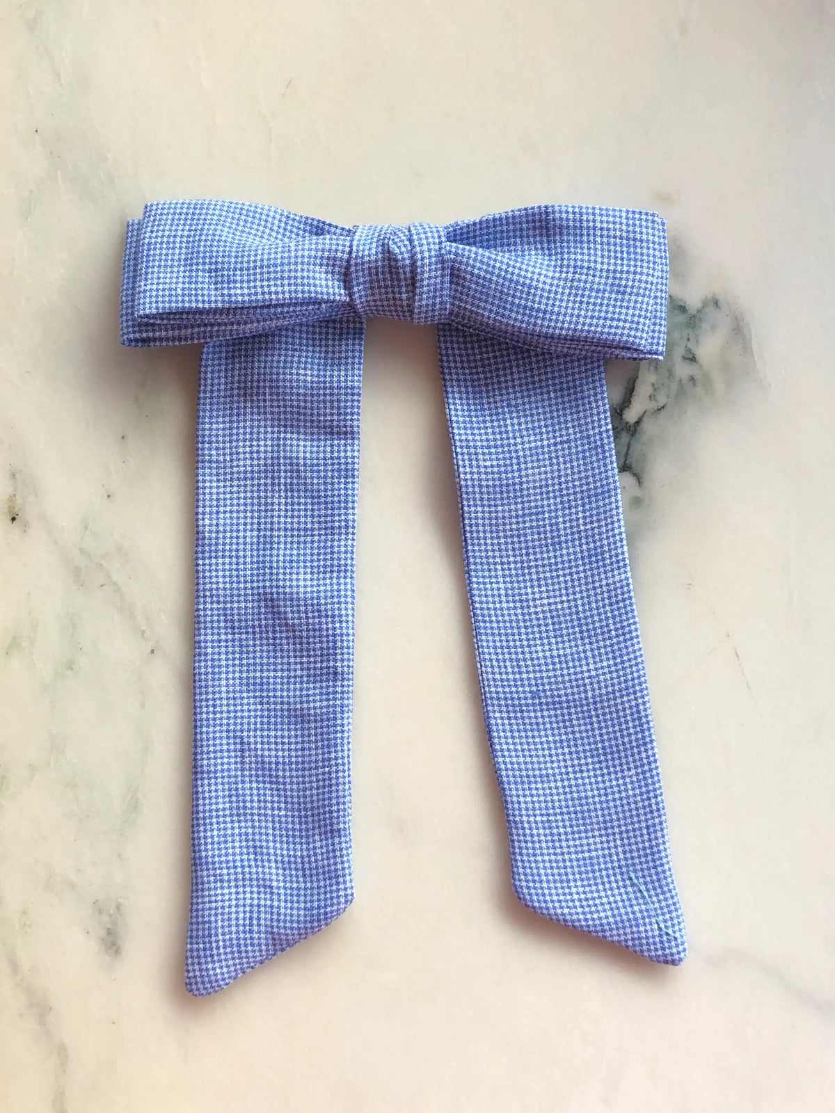 Broochella Sofia blue check linen bow brooch, a handmade designer statement accessory with an oversized bow and elegant long ribbon tails, photographed on white marble.
