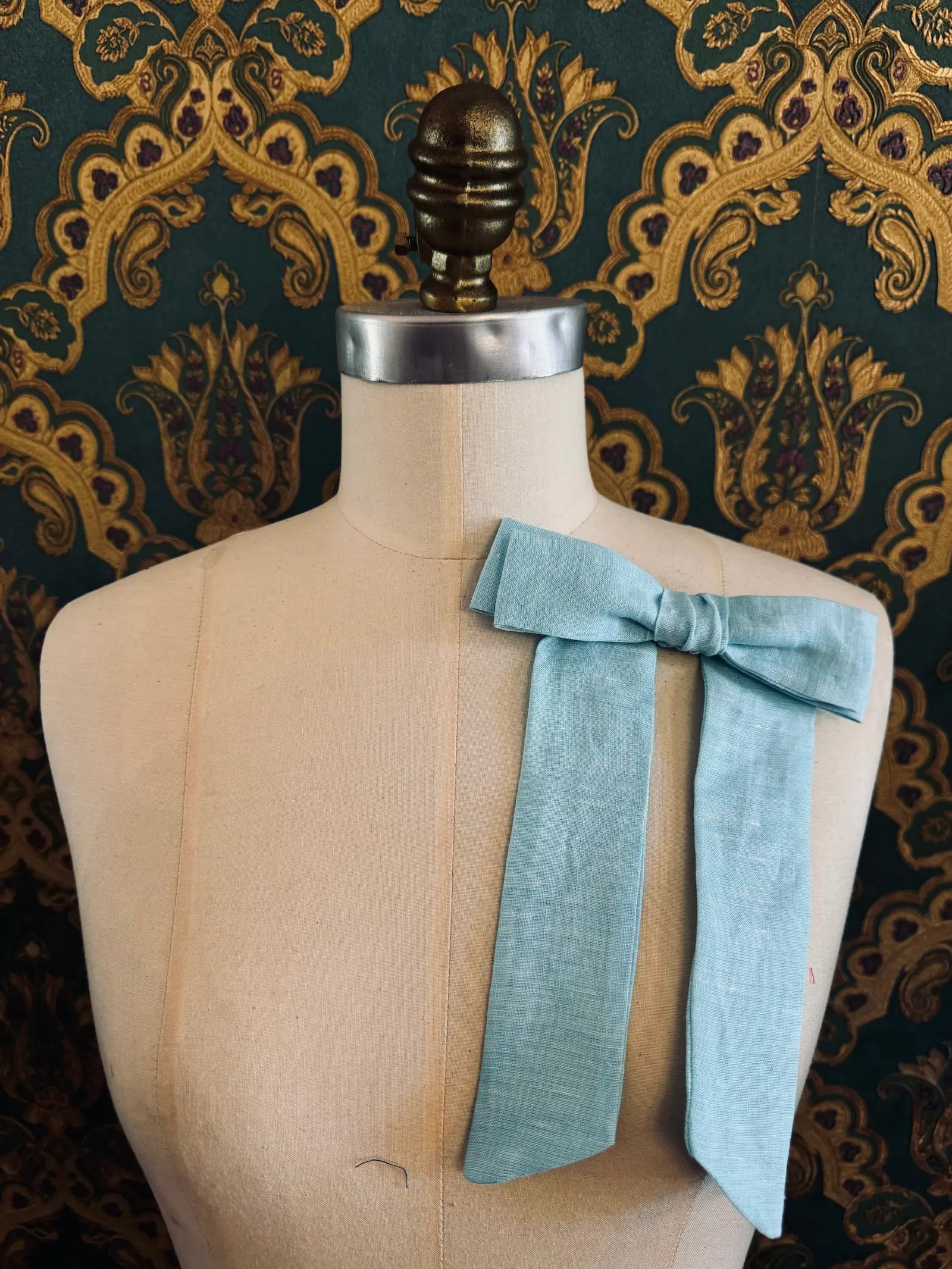 Broochella  Sofia handmade linen bow brooch in blue. Pictured on a mannequin. The background is green and gold damask wallpaper.