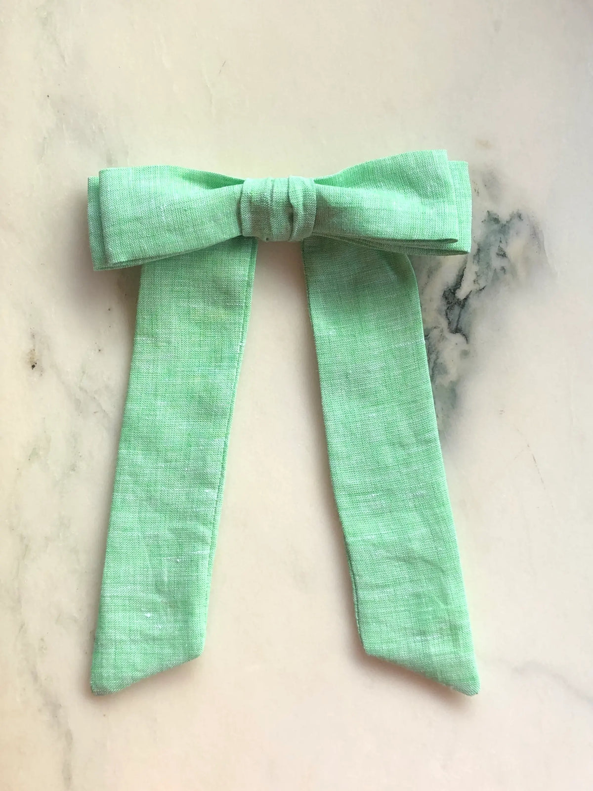 Broochella Sofia green linen bow brooch, a handmade designer statement accessory with an oversized bow and elegant long ribbon tails, photographed on white marble.