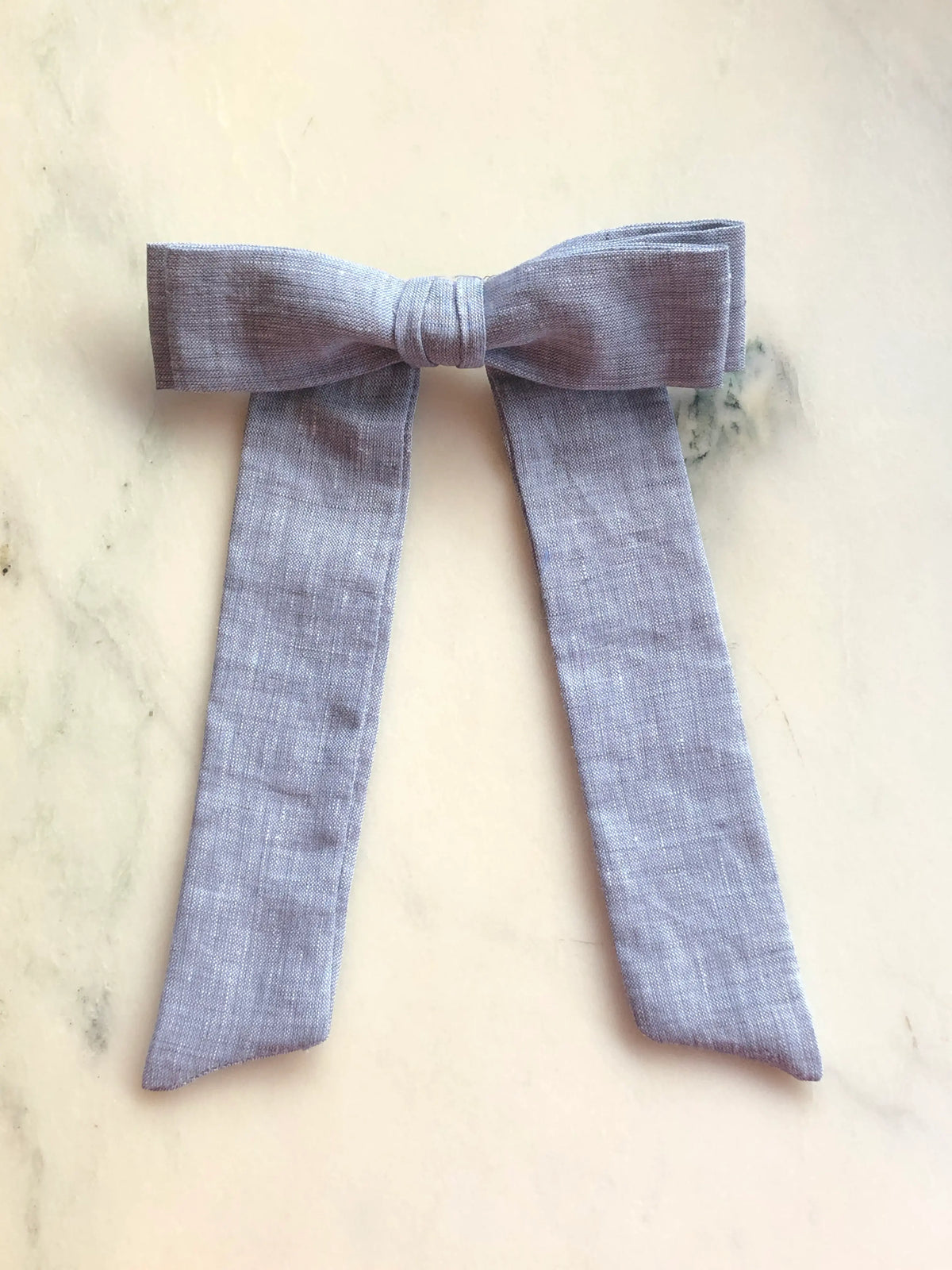 Broochella Sofia grey linen bow brooch, a handmade designer statement accessory with an oversized bow and elegant long ribbon tails, photographed on white marble.