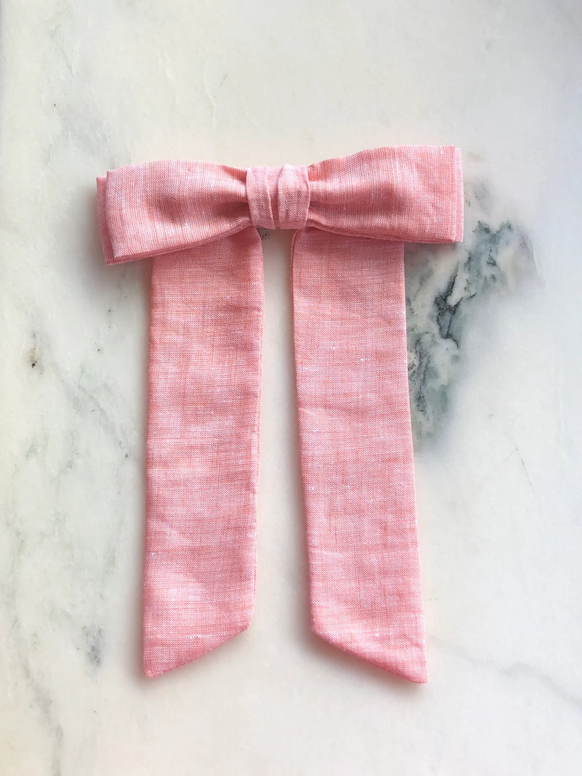 Broochella Sofia peach linen bow brooch, a handmade designer statement accessory with an oversized bow and elegant long ribbon tails, photographed on white marble.