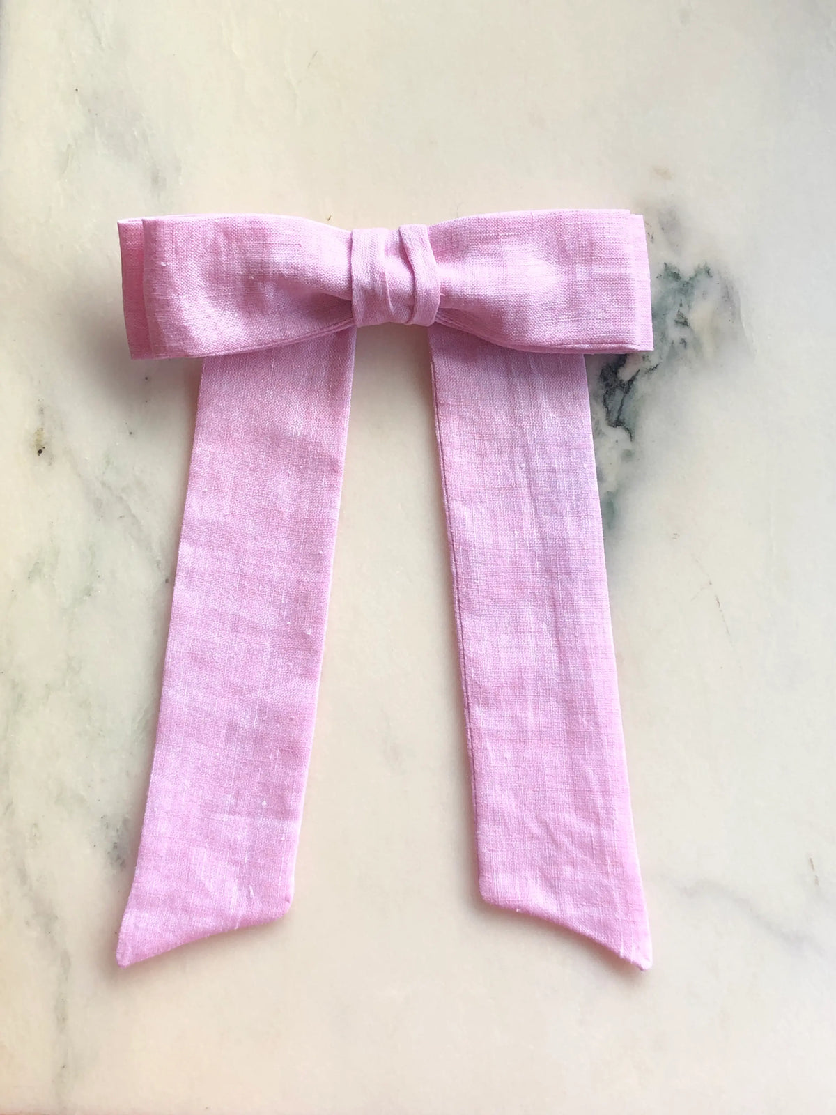 Broochella Sofia pink linen bow brooch, a handmade designer statement accessory with an oversized bow and elegant long ribbon tails, photographed on white marble.