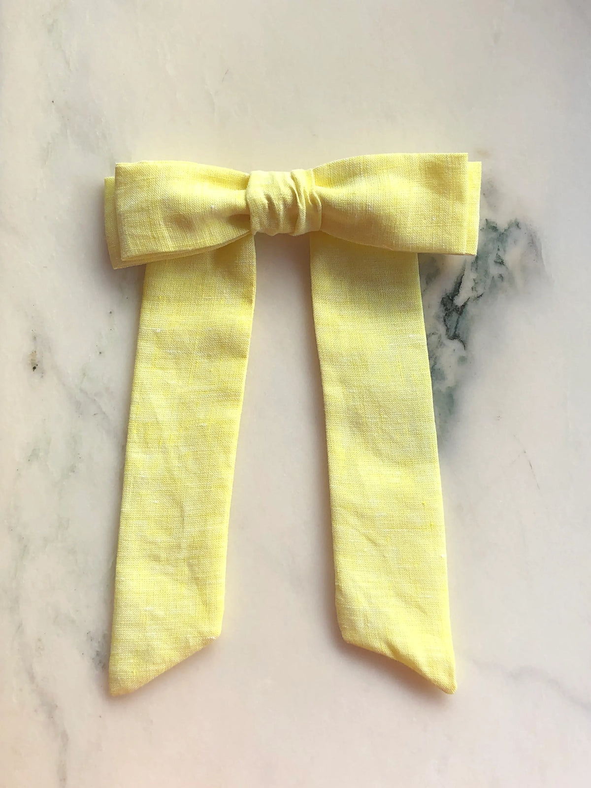 Broochella Sofia yellow linen bow brooch, a handmade designer statement accessory with an oversized bow and elegant long ribbon tails, photographed on white marble.