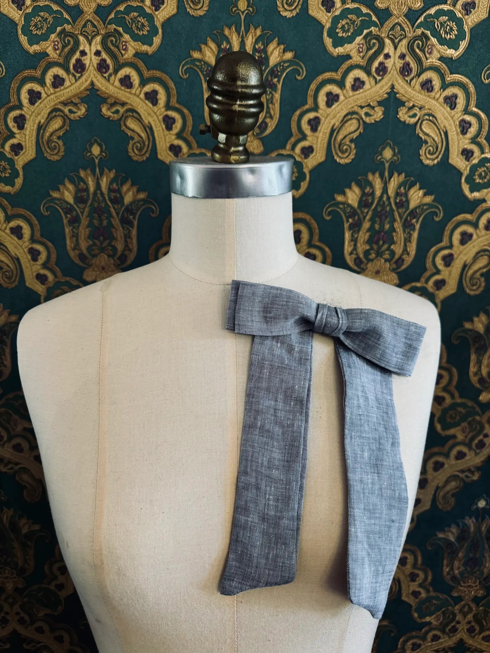 Broochella  Sofia handmade linen bow brooch in grey. Pictured on a mannequin. The background is green and gold damask wallpaper.