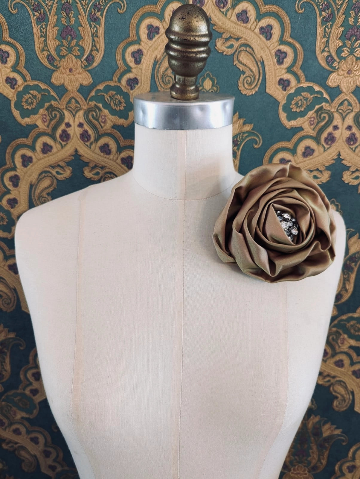 Broochella sofia bloom handmade flower brooch in olive satin. The centre is a vintage crystal brooch. The brooch is pinned to a mannequin. The background is green and gold damask wallpaper.