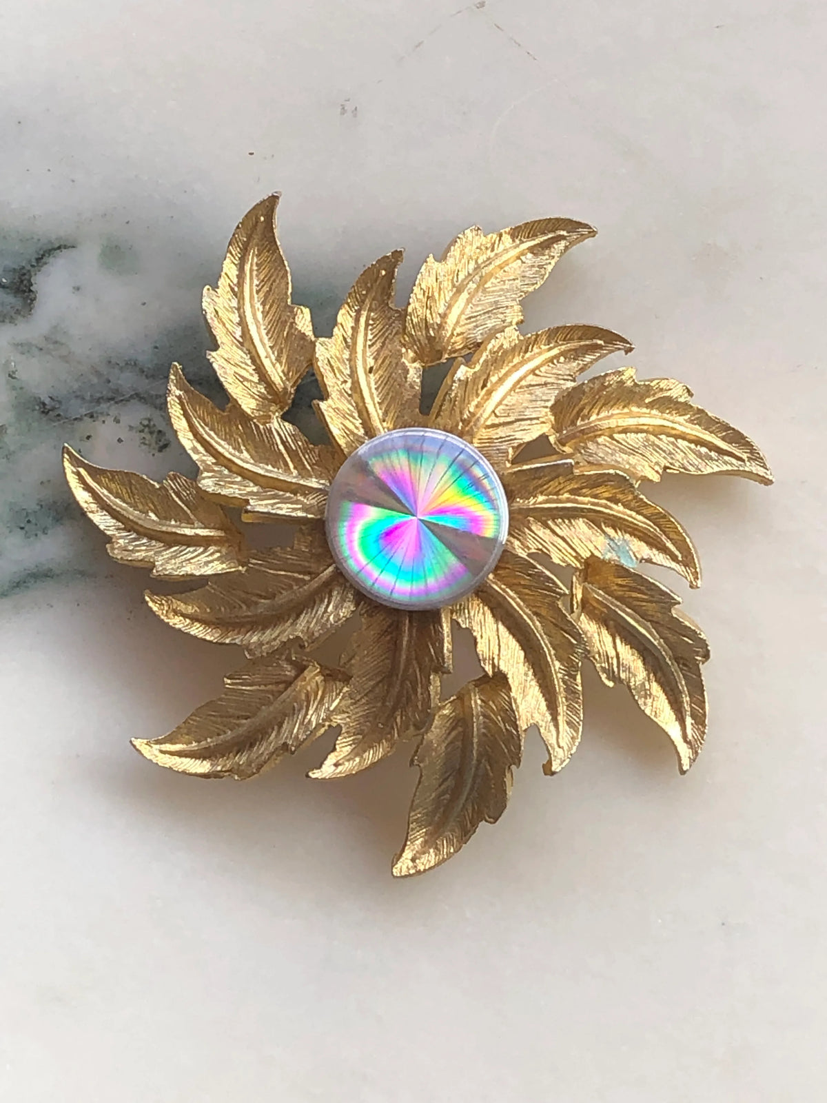 Broochella Sole d’Oro Vintage Brooch with layered textured gold-tone leaves radiating around an iridescent rainbow-effect circular centre, photographed on white marble with natural grey-green veining.