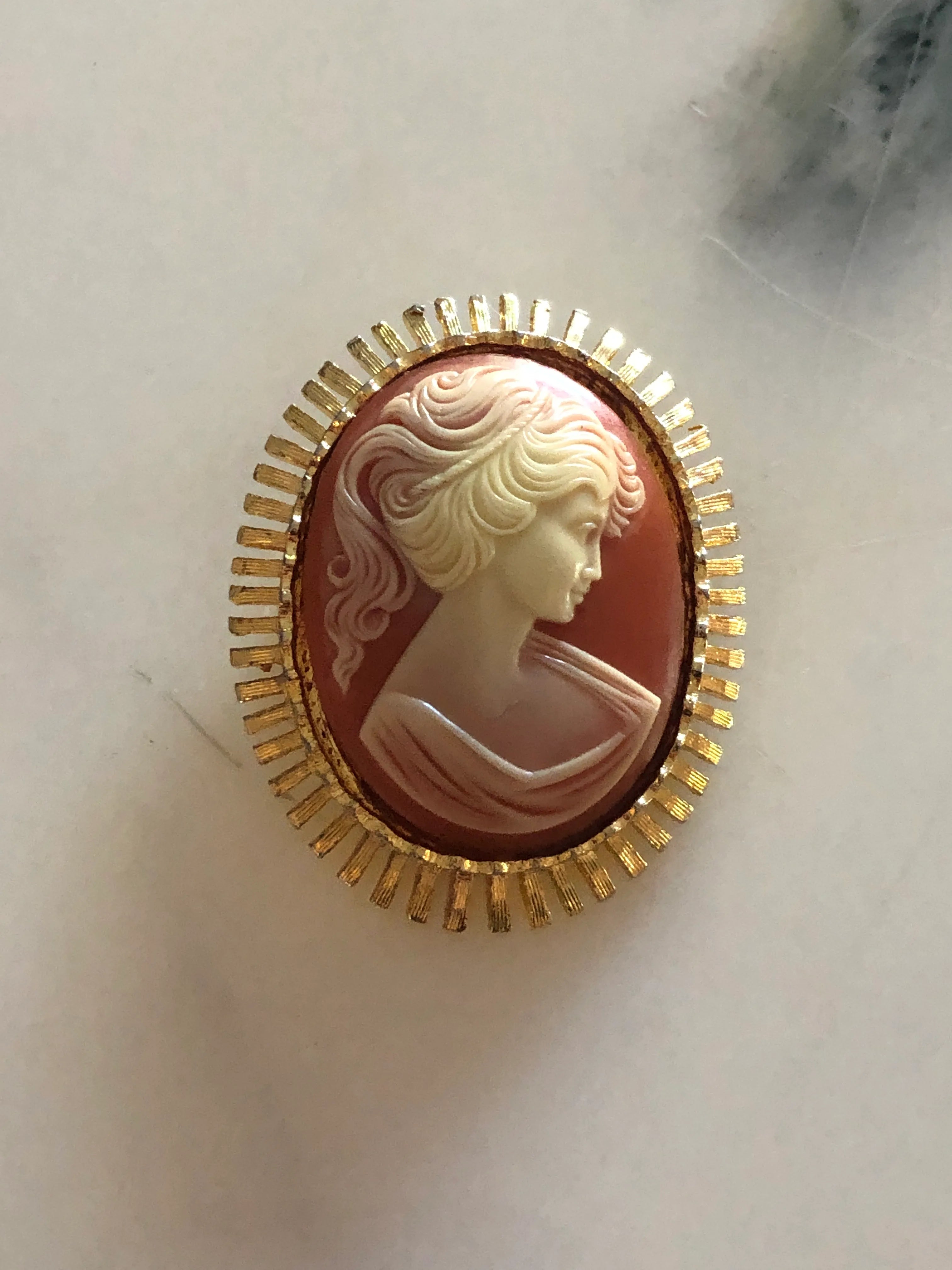 A 5cm x 4cm Broochella sole vintage cameo brooch featuring a carved cream classical portrait on a rich terracotta ground set within a gold-tone sunburst radiating bar frame, photographed on a white marble surface.