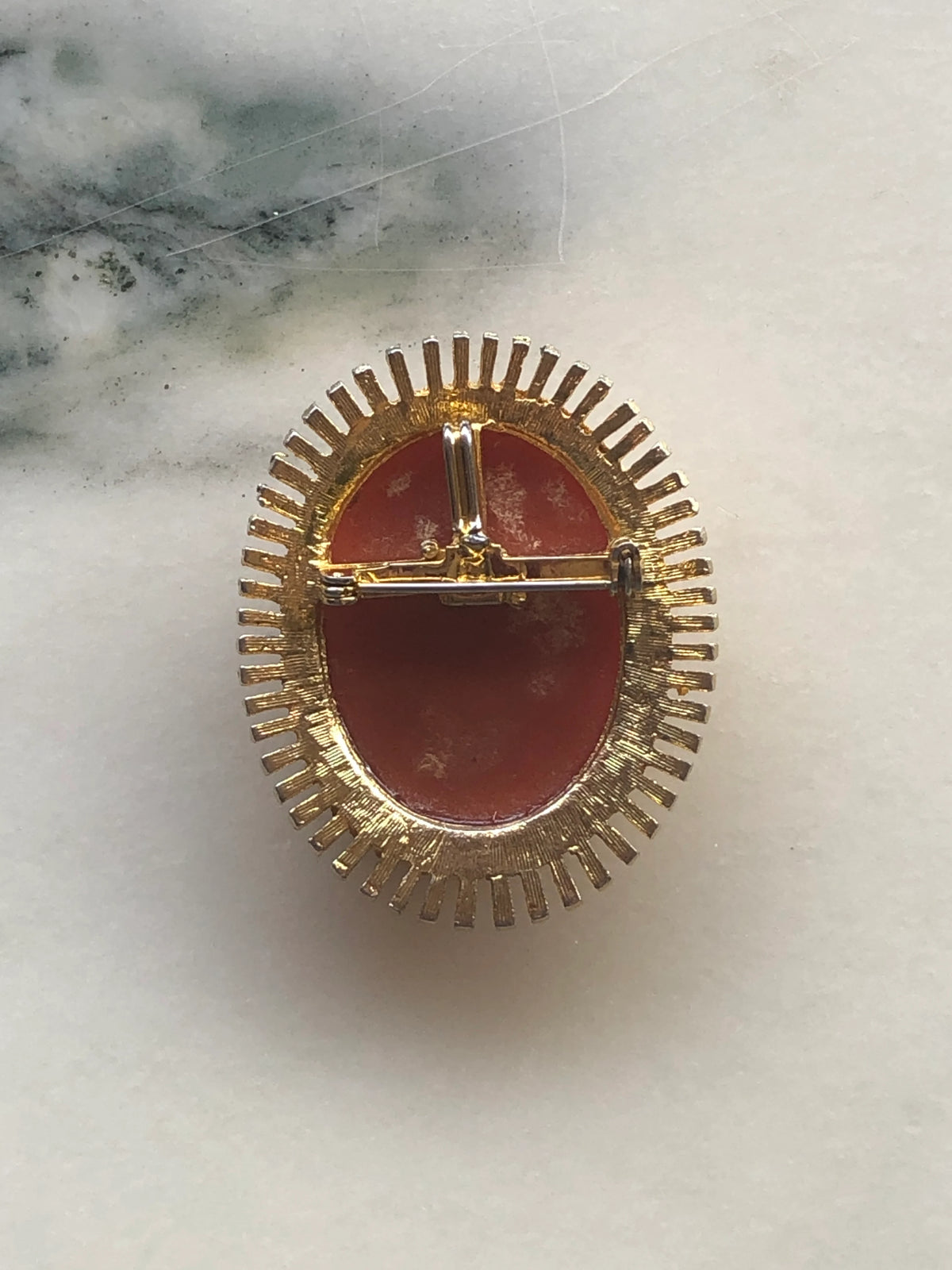 The back view of A 5cm x 4cm Broochella sole vintage cameo brooch featuring a carved cream classical portrait on a rich terracotta ground set within a gold-tone sunburst radiating bar frame, photographed on a white marble surface.