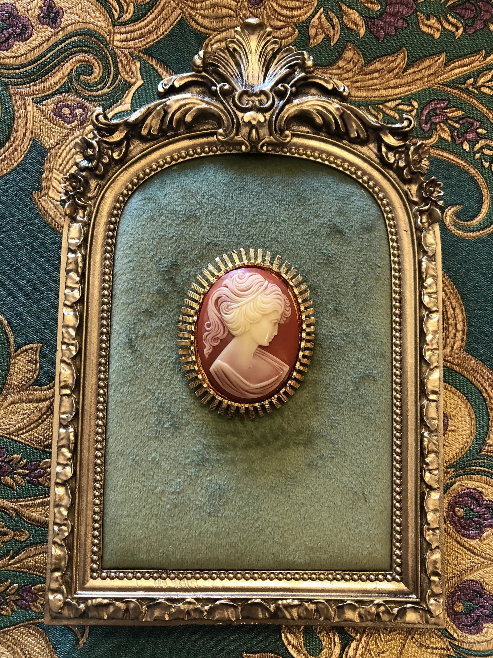 A 5cm x 4cm Broochella Sole vintage cameo brooch featuring a carved cream classical portrait on a rich terracotta ground set within a gold-tone sunburst radiating bar frame. It is photographed on a green velvet tray, with an ornate gold frame. The background is green and gold damask wallpaper. 