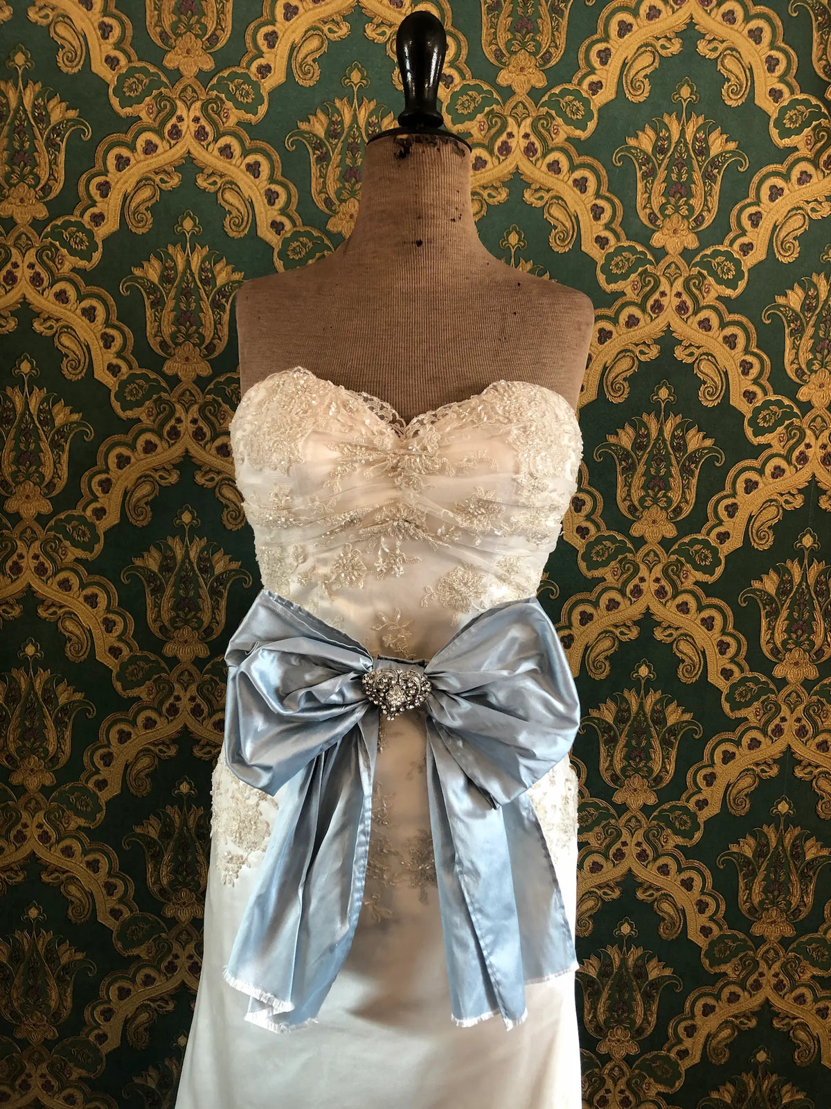 Broochella Something Blue Silk Bow in powder blue silk with a heart-shaped crystal brooch centre, styled at the waist of an ivory lace wedding dress on a mannequin against ornate green and gold wallpaper.