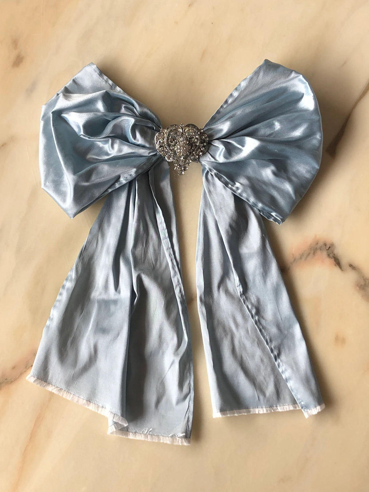 Broochella Something Blue Silk Bow in powder blue with a heart-shaped crystal brooch centre and long flowing silk tails, photographed on a white marble background.