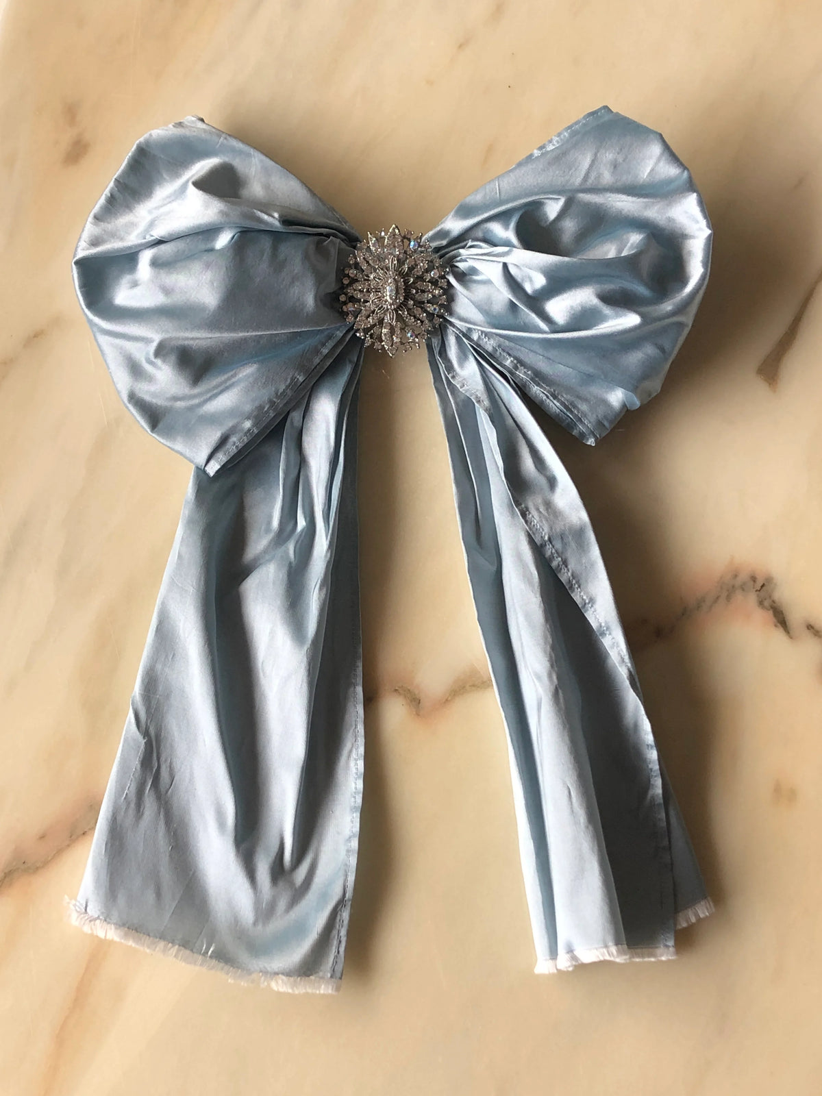 Broochella Something Blue Silk Bow in powder blue with an ornate oval crystal brooch centre and long flowing silk tails, photographed on a white marble background.