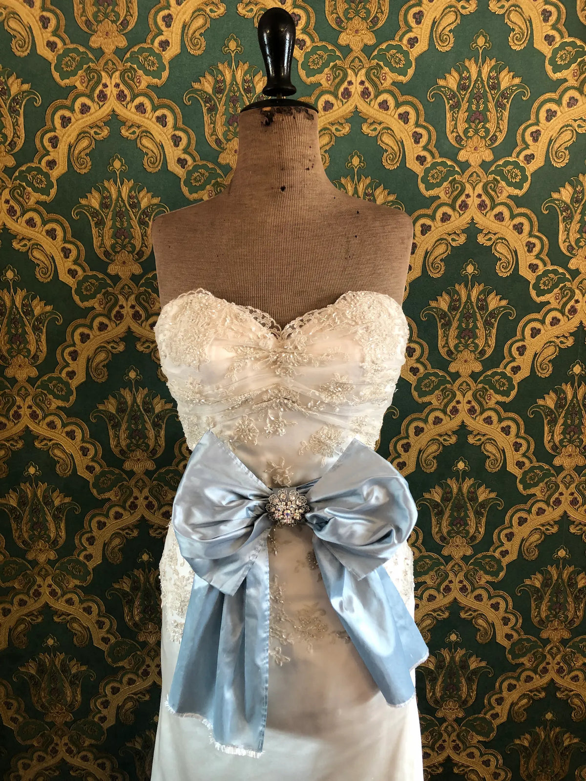 Broochella Something Blue Silk Bow in powder blue silk with a sparkling round crystal brooch centre, styled at the waist of an ivory lace wedding dress on a mannequin against ornate green and gold wallpaper.