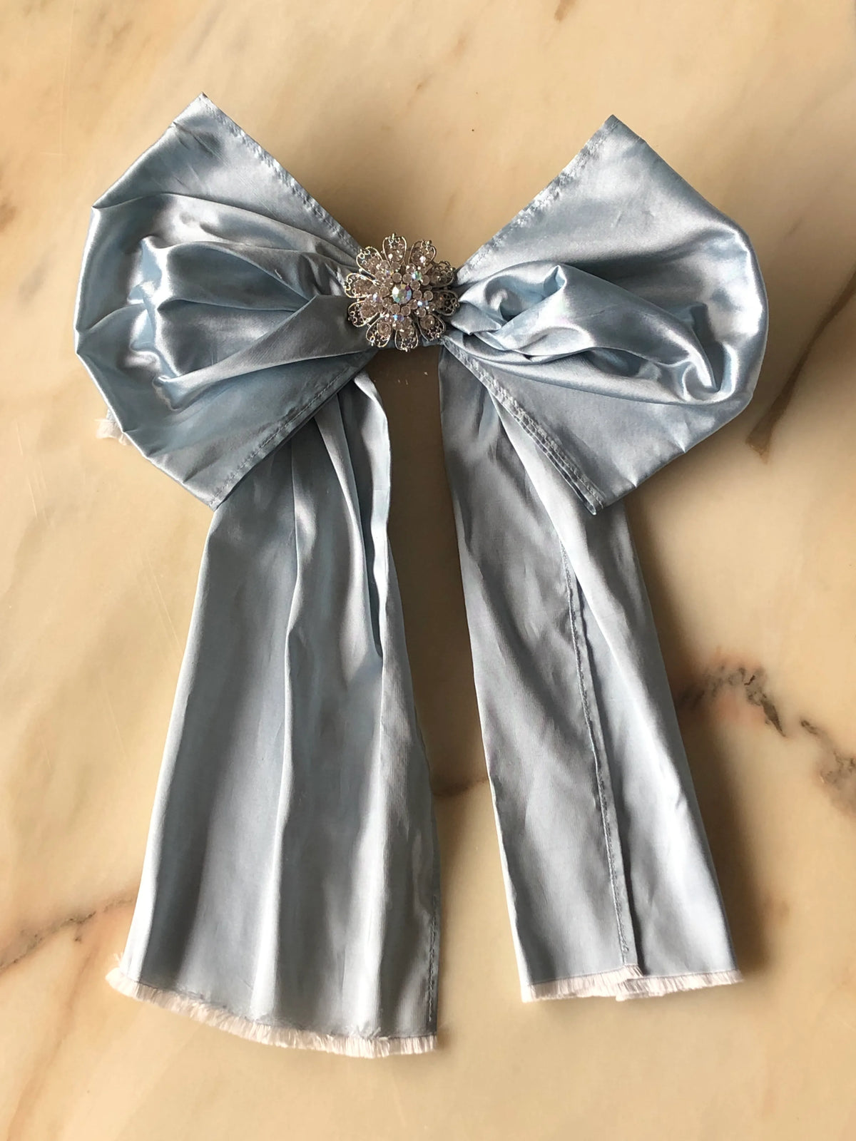 Broochella Something Blue Silk Bow in powder blue with a sparkling round crystal brooch centre and long flowing silk tails, photographed on a white marble background.