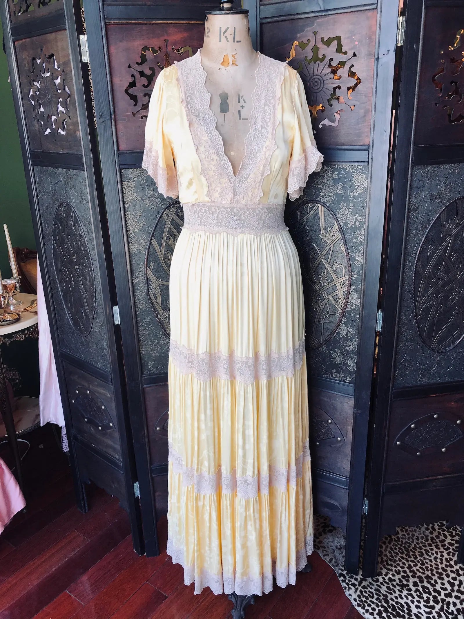 Broochella Spell maxi dress size small. Soft butter / cream 100% Viscose fabric, with deep V neckline and flutter sleeve. It has Lace-trim detailing and a pleated maxi skirt with beige lace panel inserts. It is pictured on a mannequin. The background is an ornate, dark wood, vintage room divider. 
