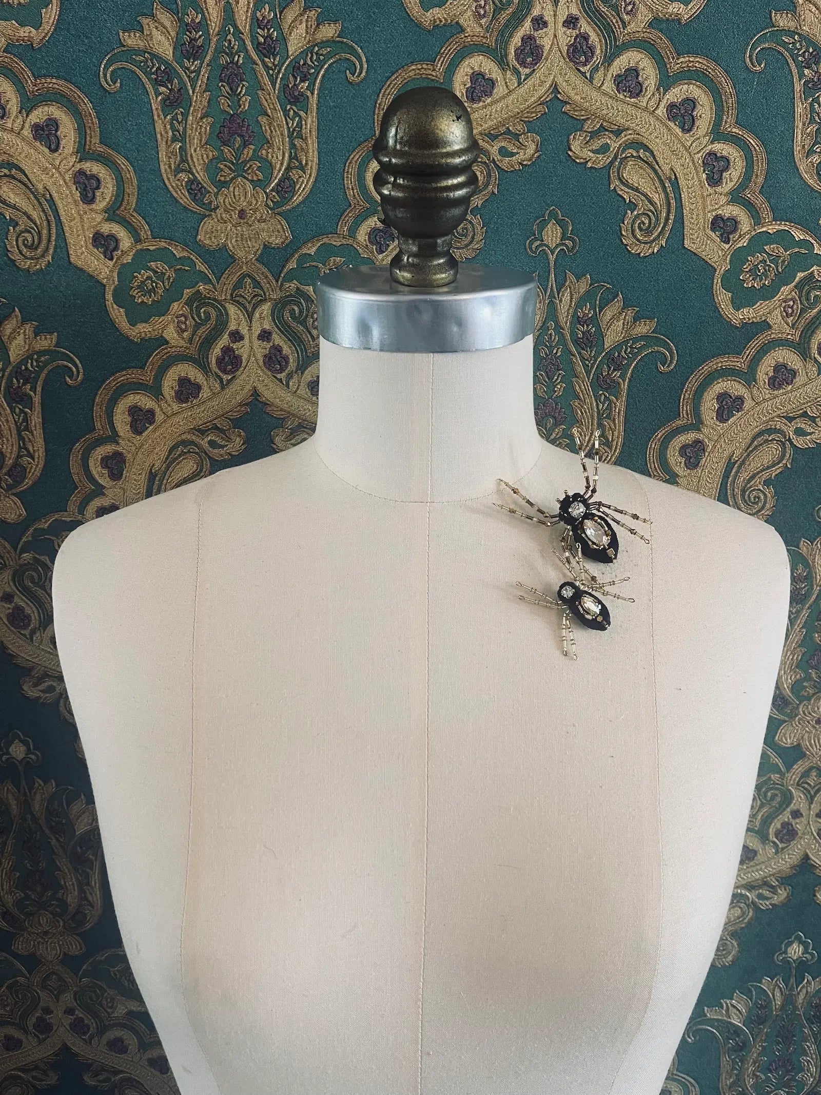 Broochella black and gold hand beaded spider brooch set. Pictured on a mannequin. The background is green and gold damask wallpaper