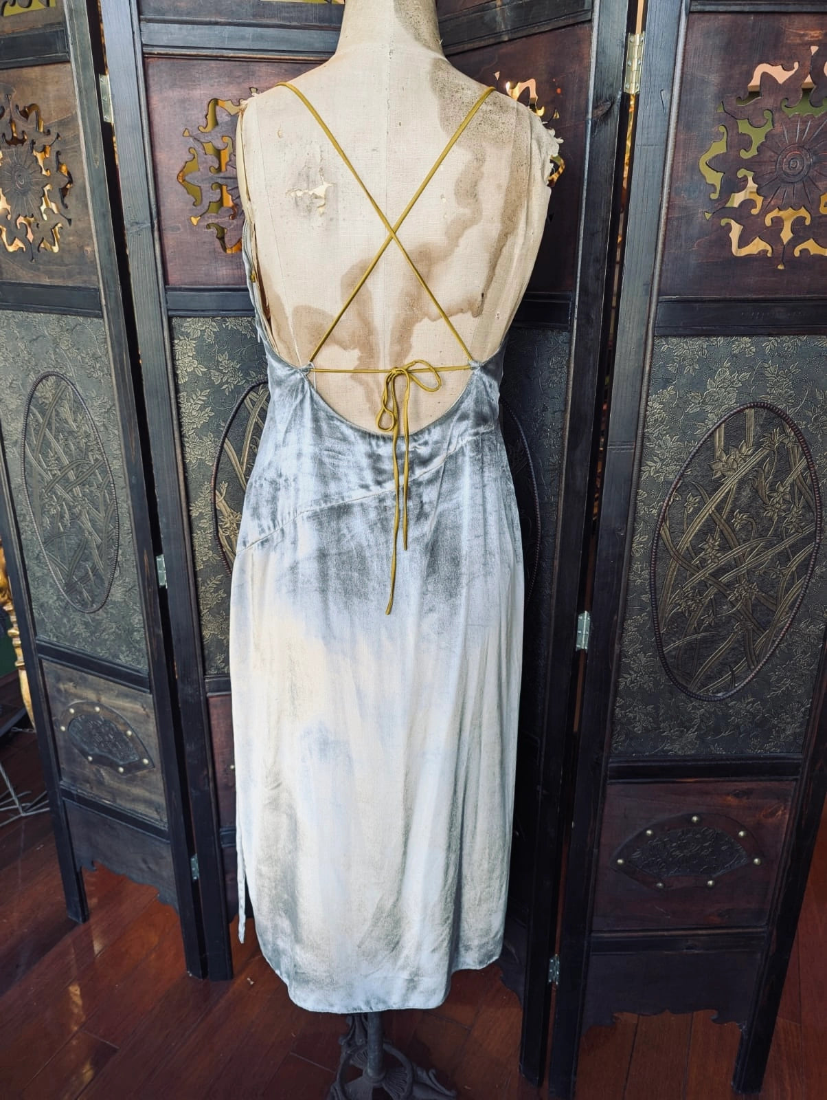 The back view of Broochella Ssone silver velvet slip dress, with gold straps. It is pictured on a mannequin. The background is an ornate, dark wood room divider. 