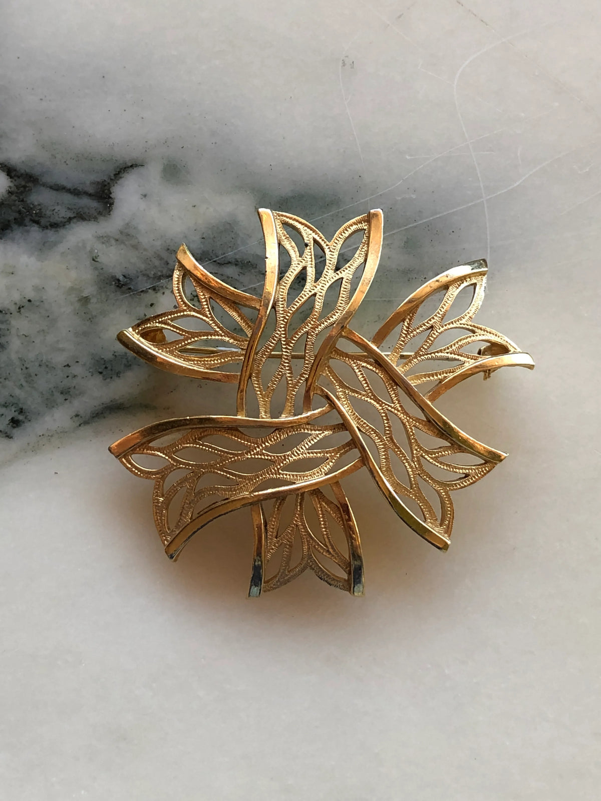 Broochella Stella Dorata Trifari Vintage Brooch Pin front view featuring sculptural overlapping gold-tone openwork leaves with delicate textured detailing, photographed on white marble with soft grey-green veining.