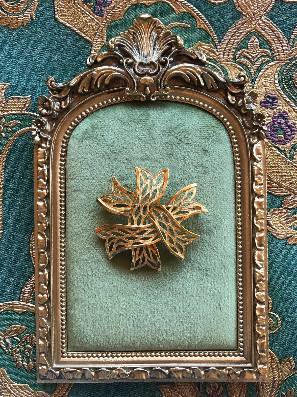 Broochella Stella Dorata Trifari Vintage Brooch Pin, an intricate gold-tone openwork starburst of overlapping leaf forms, displayed on sage green velvet in an ornate antique gold frame against green and gold patterned fabric.
