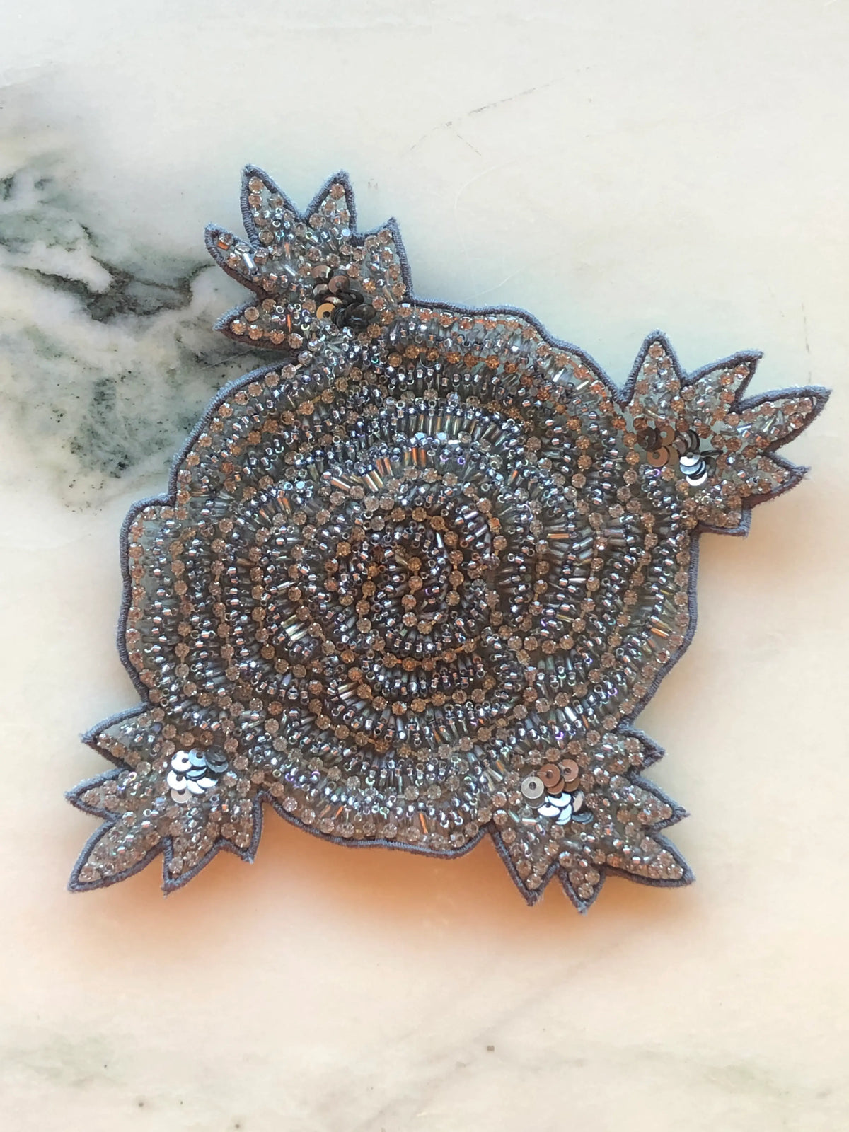 Broochella Stella Hand-Beaded Brooch featuring an ornate floral design intricately embellished with silver, champagne and metallic beads, crystals and sequins on a cream marble background.