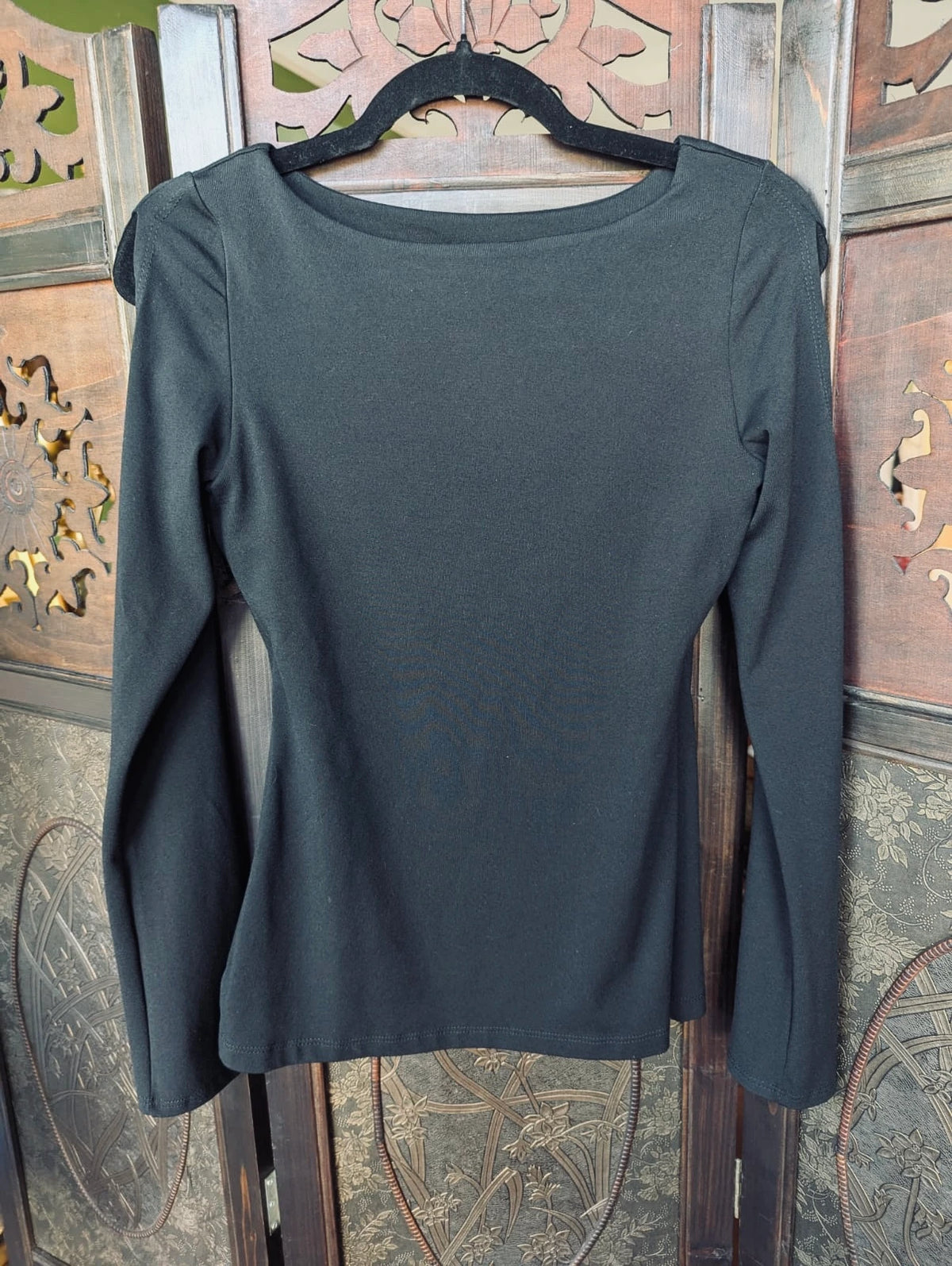 A Broochella Susana Monaco black  long-sleeve top with a boat neckline, hanging on a black hanger against a decorative wooden screen.