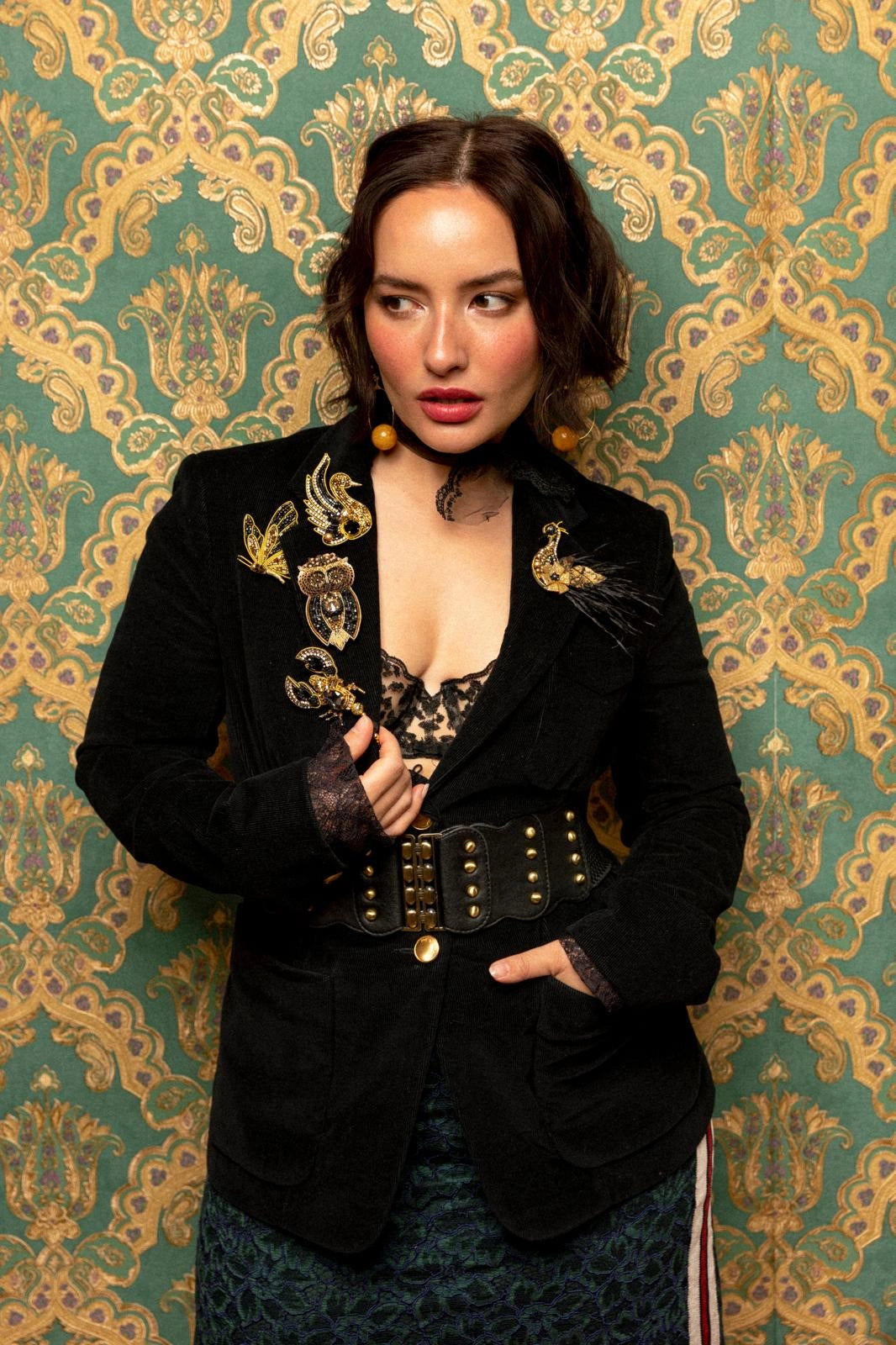 A woman posing in front of ornate green and gold damask wallpaper, wearing a black velvet blazer with gold embroidery, a wide black belt with gold studs, and a patterned skirt. She is wearing the Broochella gold and black hand beaded swan brooch