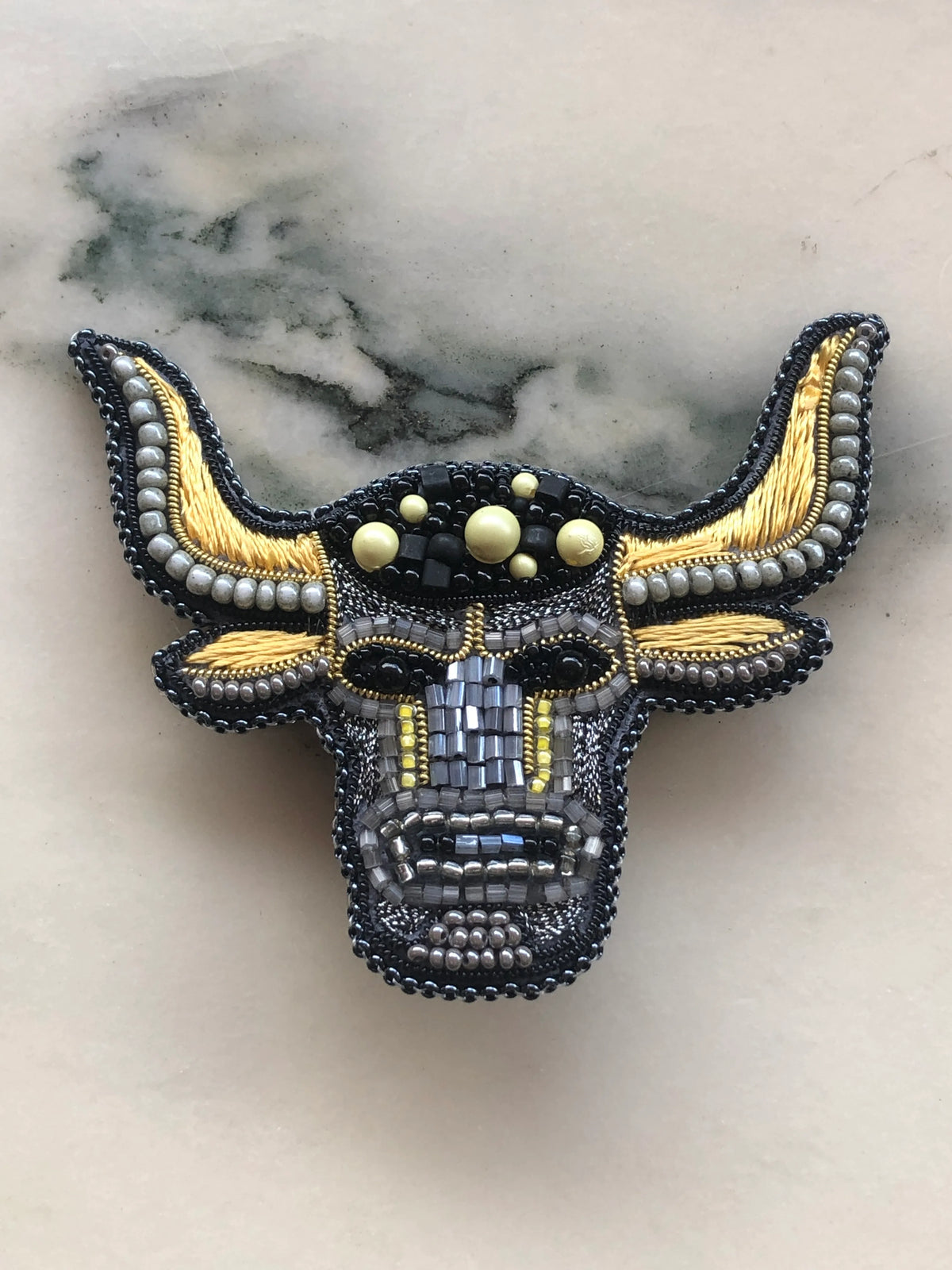 A Broochella Taurus Zodiac handcrafted bull head brooch featuring intricate black and grey beading, gold metallic accents, and cream-coloured decorative beads, displayed on a light marble surface.