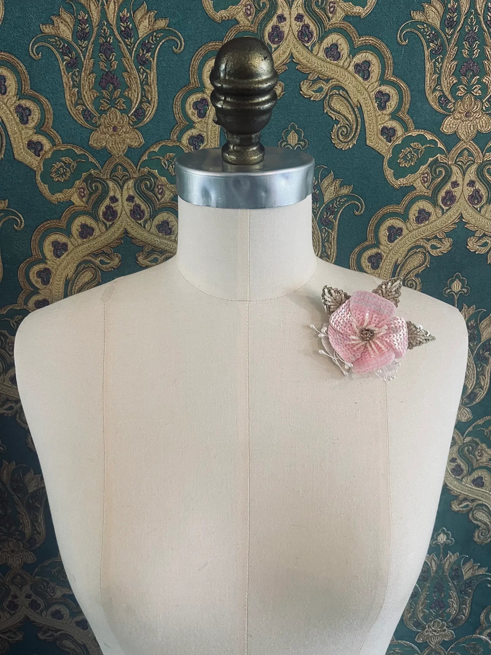 Broochella Tesorino Ingioiellato hand beaded pink, white and silver flower brooch. It is pinned to a mannequin. The background is green and gold damask wallpaper.