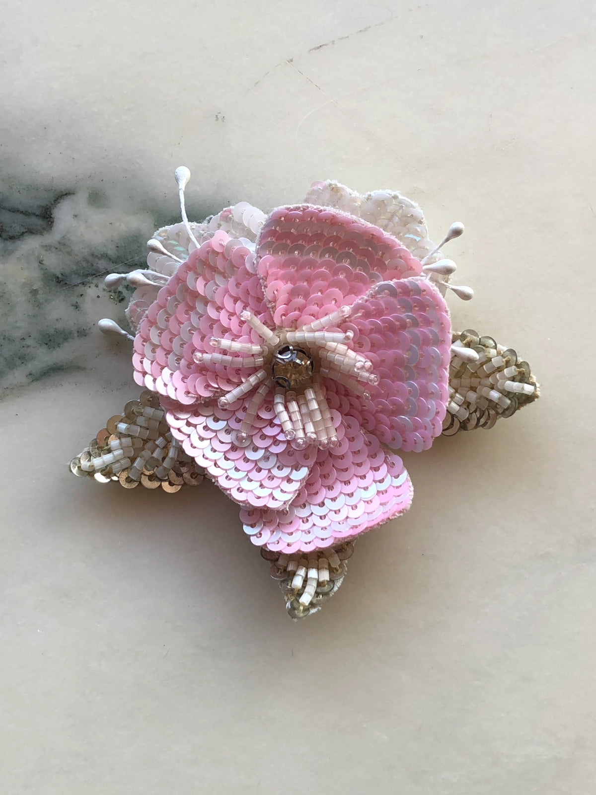 Broochella Tesorino Sequinned Flower Brooch Pin with layered pink sequinned petals, ivory beading and crystal details, photographed on a white marble background with soft grey veining.