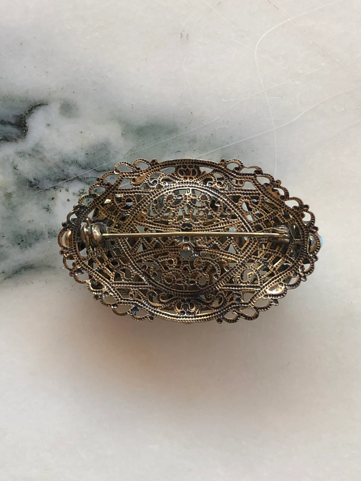 The back view of Broochella tesoro azzurro czech filigree vintage oval brooch with a turquoise-blue central stone and small light blue stones around it, set in gold-toned metal. It is displayed on a white marble surface