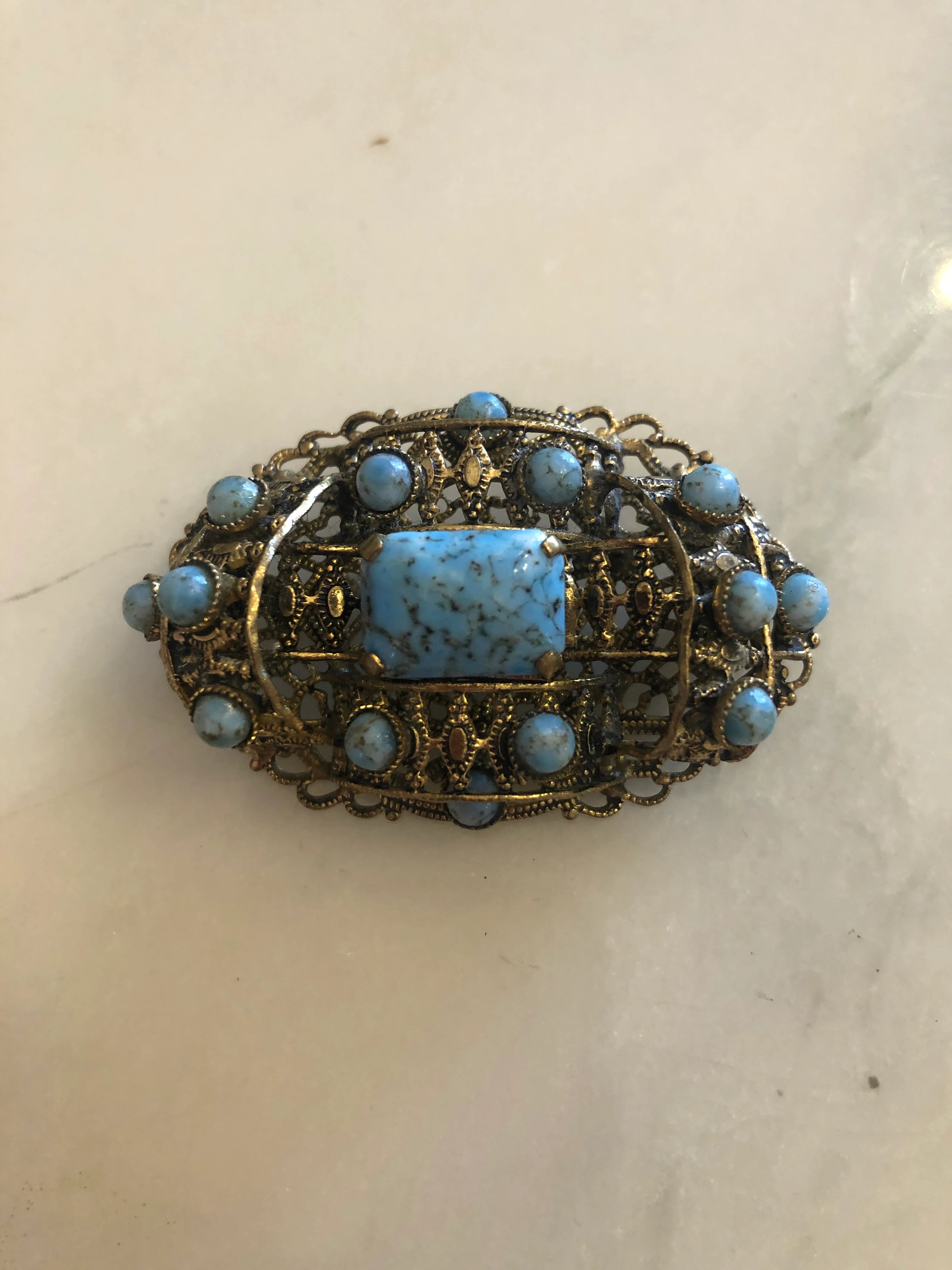 Broochella tesoro azzurro czech filigree vintage oval brooch with a turquoise-blue central stone and small light blue stones around it, set in gold-toned metal. It is displayed on a white marble surface