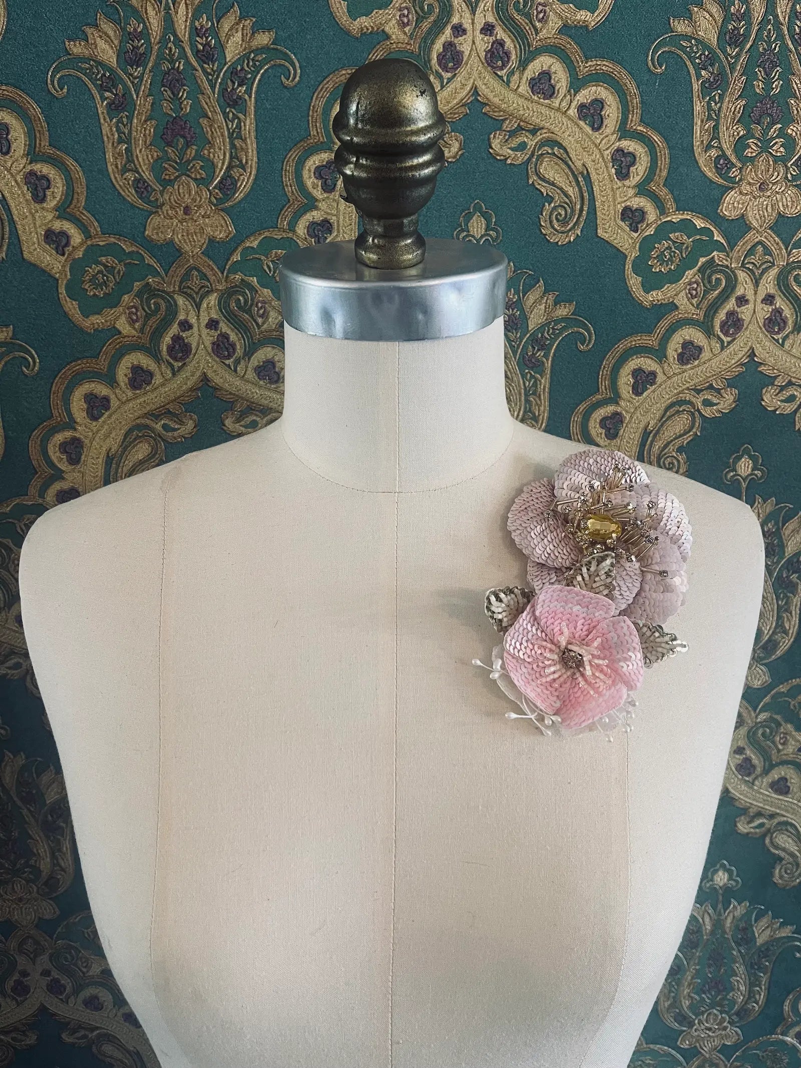 Broochella tesoro ingioiellato handmade sequin pink and gold flower brooch. Pictured on a mannequin. The background is green and gold damask wallpaper