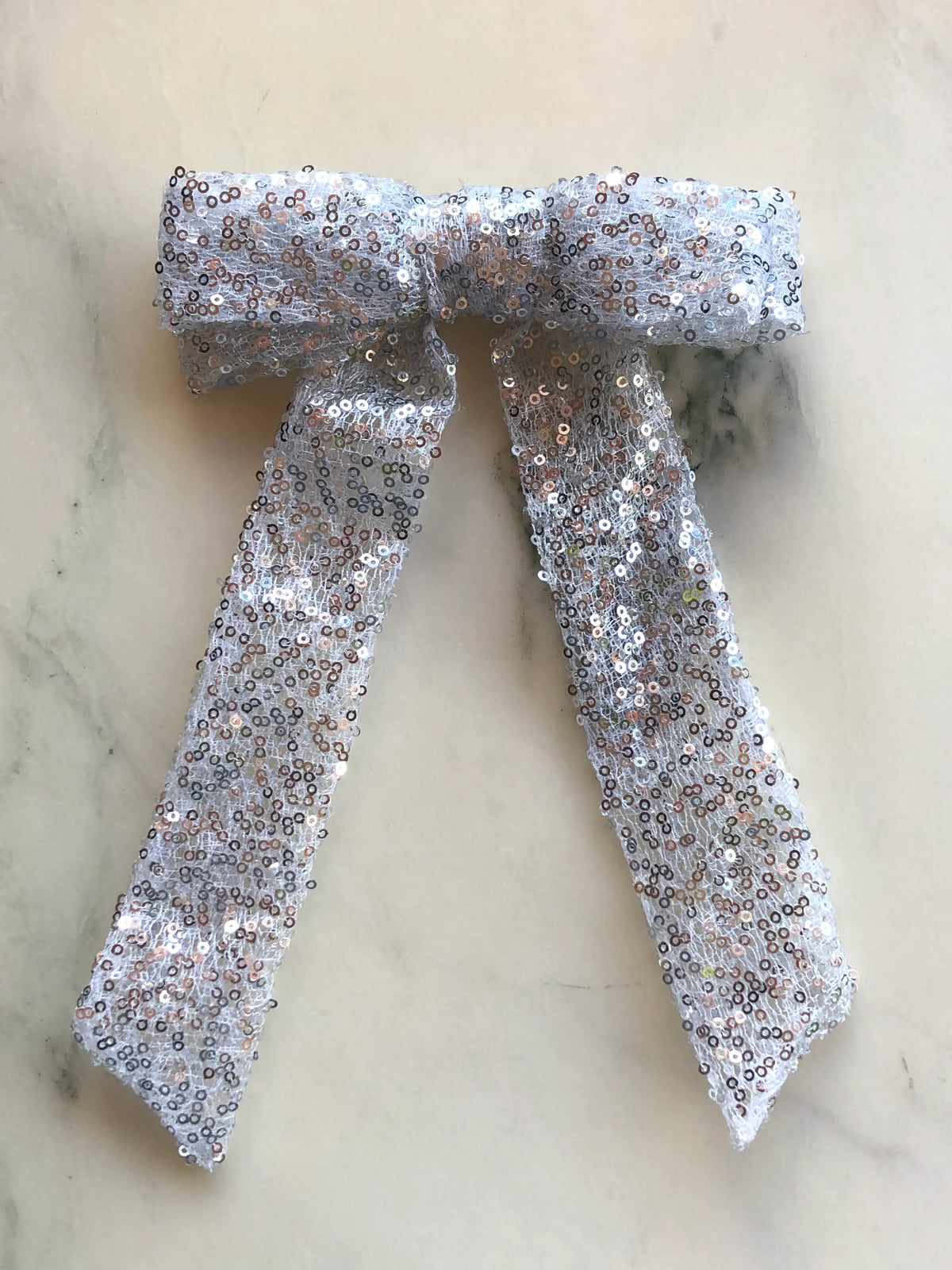Broochella The Stardust Sequin Bow Brooch in shimmering silver sequins with long ribbon tails, photographed on a white marble background.
