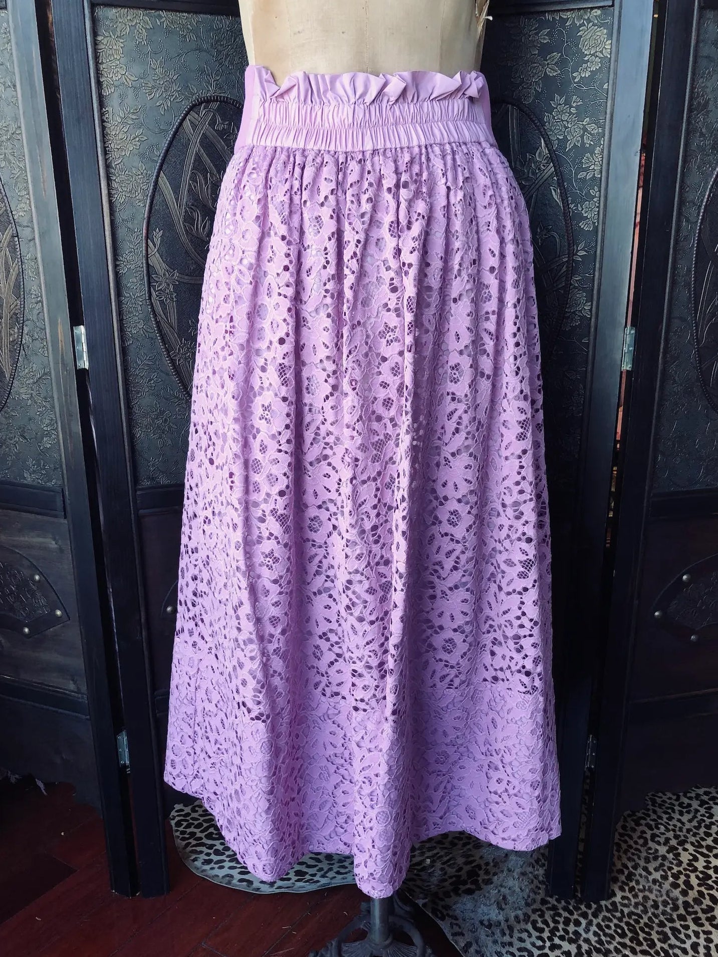 A Broochella Tibi lavender purple eyelet lace midi skirt with a ruffled waistband, displayed on a dress form mannequin in front of a dark wooden folding screen.