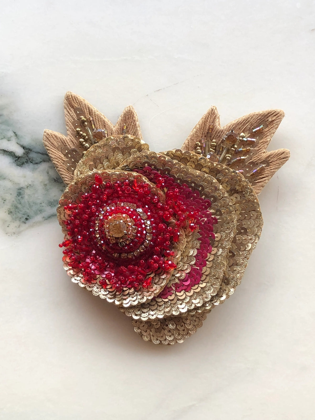 Broochella Tonina Tesorina Embellished Brooch Pin featuring layered gold and deep red sequins, red beadwork, sparkling crystal details and embroidered gold leaves, photographed on a white marble background with soft grey-green veining.
