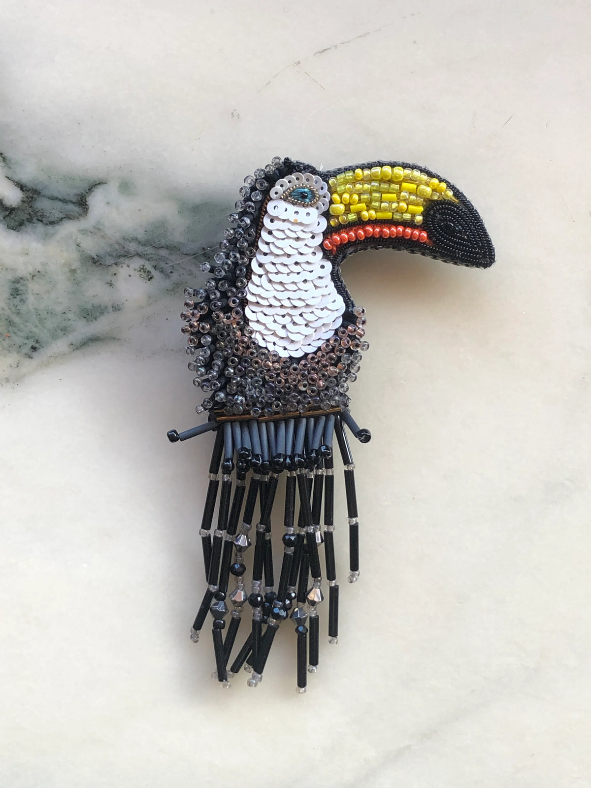 Broochella Toucan Bird Brooch hand embellished with black, white, yellow and orange beads and sequins with beaded fringe, photographed on white marble.