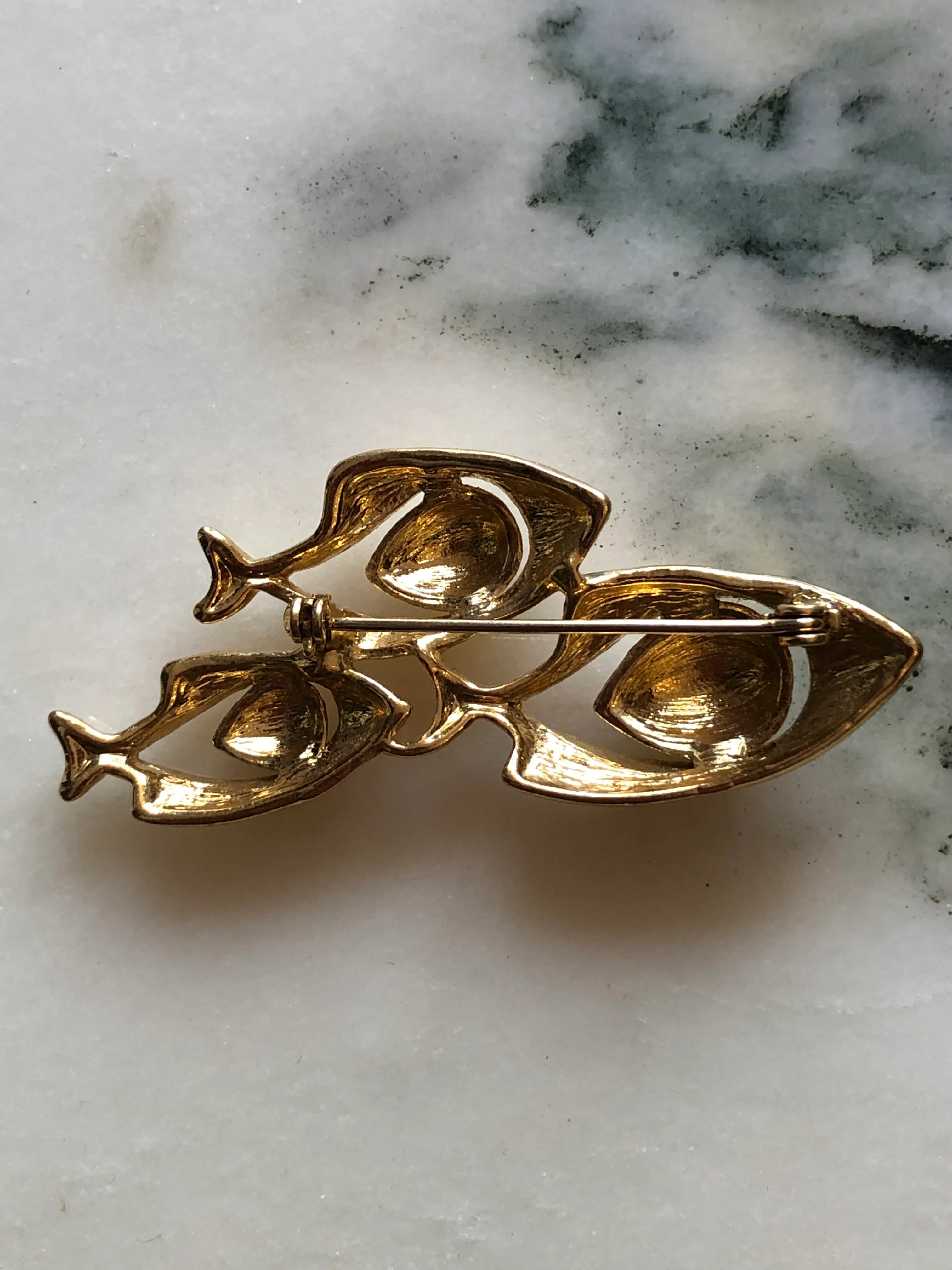 The back view of Broochella Tre Pesci vintage gold-tone brooch featuring three swimming fish with smooth ivory enamel bodies. The background is white marble.