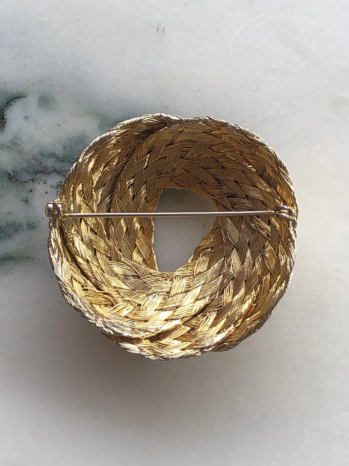 Broochella Treccia d’Oro vintage brooch pin shown from the back, revealing the detailed woven gold-tone construction and classic pin fastening, photographed on white marble with soft green veining.