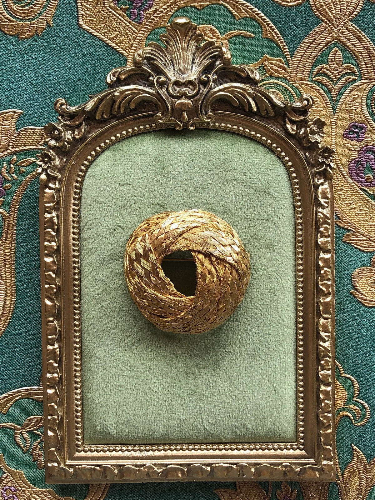 Broochella Treccia d’Oro vintage gold-tone woven brooch pin displayed on sage green velvet in an ornate antique-style gold frame, set against richly embroidered green and gold fabric.