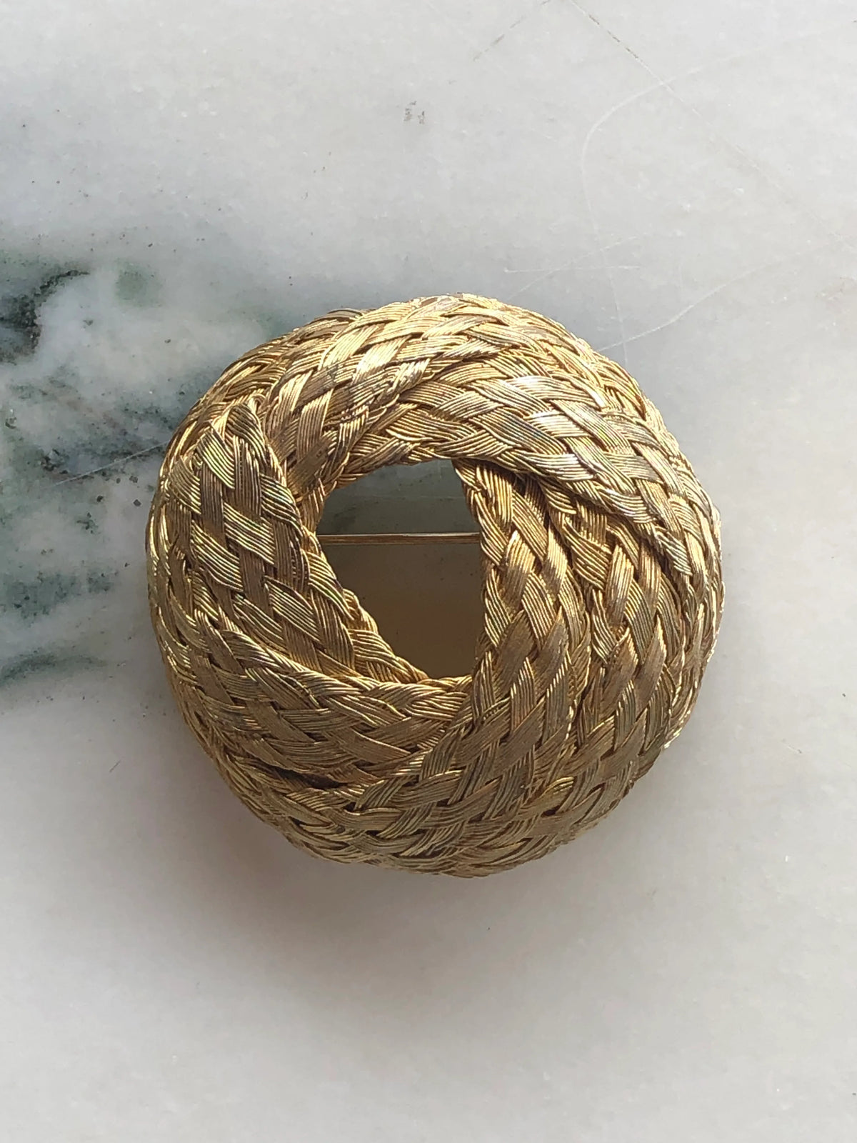 Broochella Treccia d’Oro vintage brooch pin featuring an intricate woven gold-tone circular knot design, photographed on white marble with soft green veining.