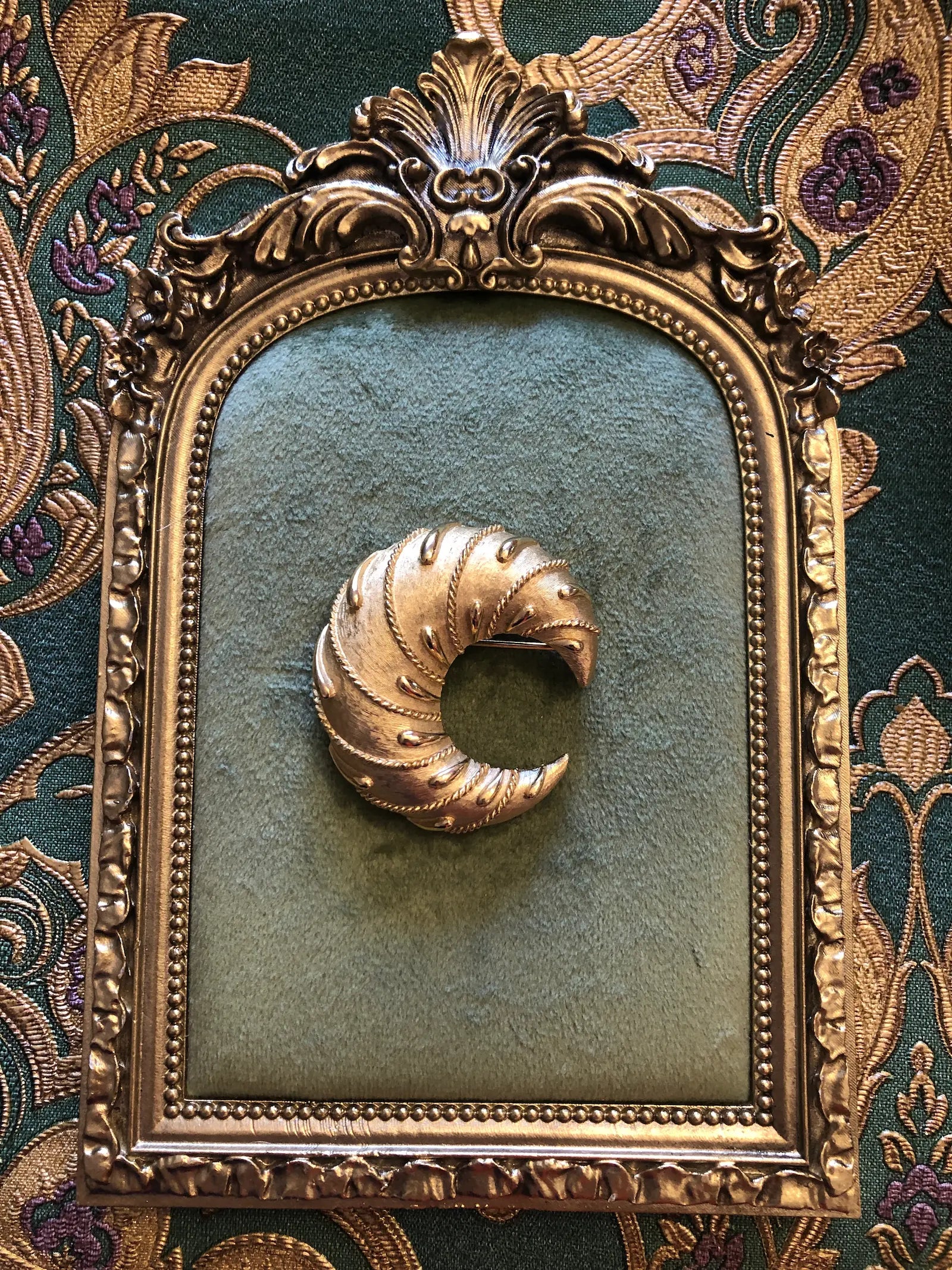 Broochella vintage trifari cornetto gold tone metal, crescent shaped brooch. It is pictured on a green velvet tray with a gold frame. The background is green and gold damask wallpaper.