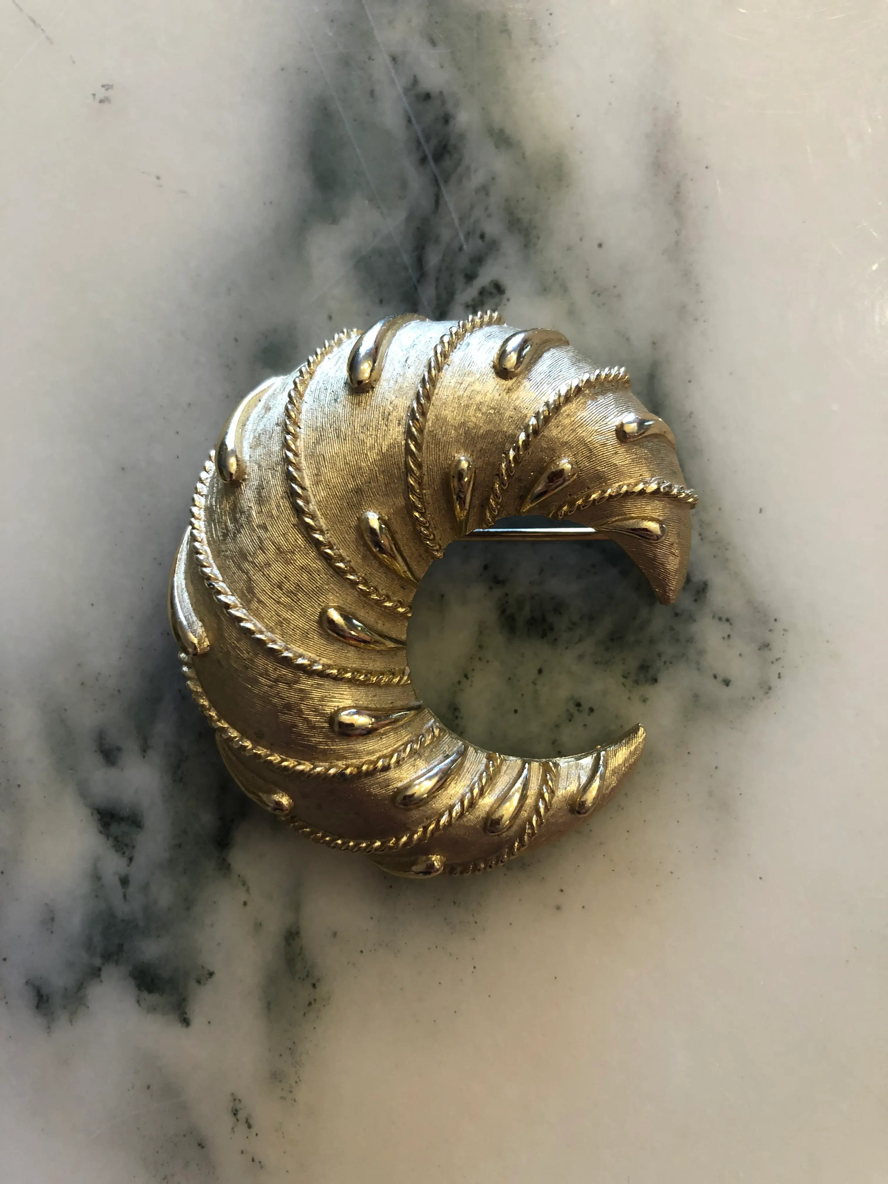 Broochella vintage trifari cornetto gold tone metal, crescent shaped brooch. The background is white marble