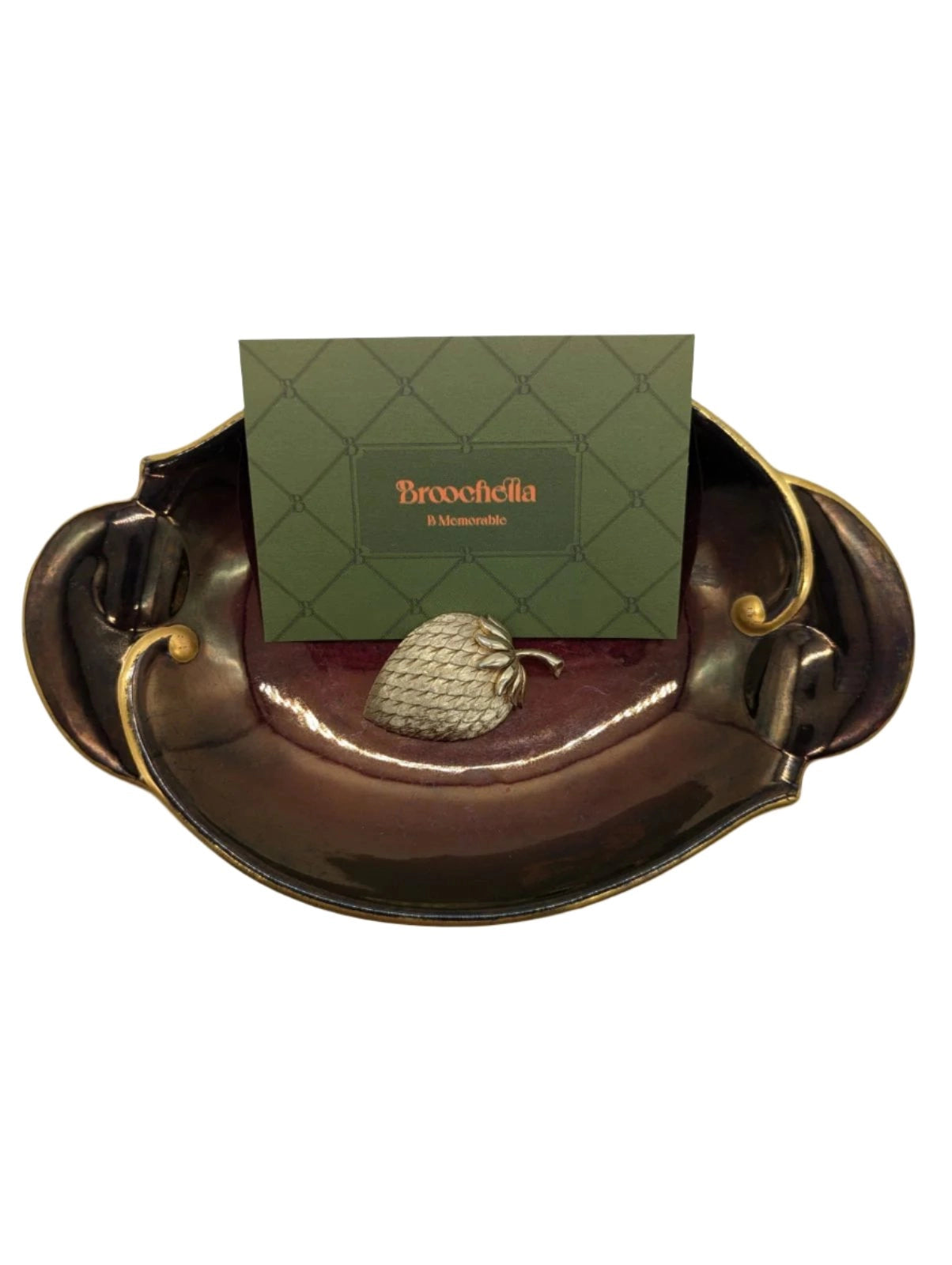 Broochella Trifari vintage gold tone metal figural strawberry brooch. Pictured on a burgundy and gold wembley ware tray. There is a green  broochella business card behind the brooch. There is a white background