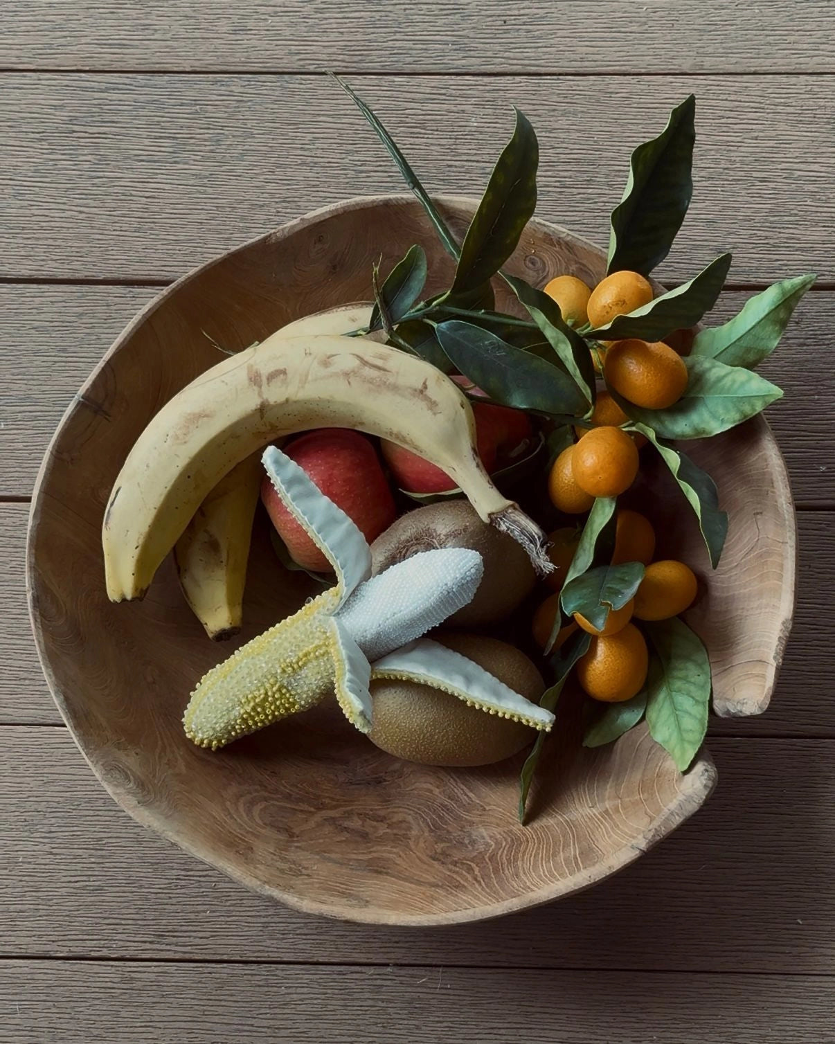A rustic wooden bowl filled with assorted fruits, including a banana, small orange fruits on branches with green leaves, kiwi, and red apples, set on a wooden surface. Place inside the bowl is the Broochella Tropicana hand beaded banana brooch
