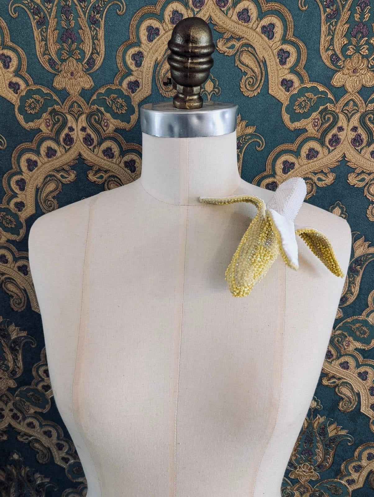 Broochella Tropicana hand beaded 3D figural banana brooch. It is pinned to a mannequin. The background is green and gold damask wallpaper.