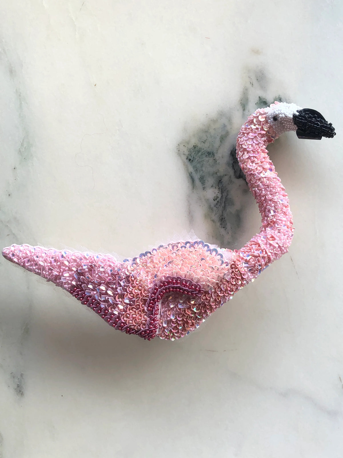 Broochella Tropicana Flamingo Brooch Pin, an intricately hand-beaded pink flamingo statement brooch embellished with shimmering sequins and beads, with a curved neck and black beak, photographed on white marble with soft grey-green veining.
